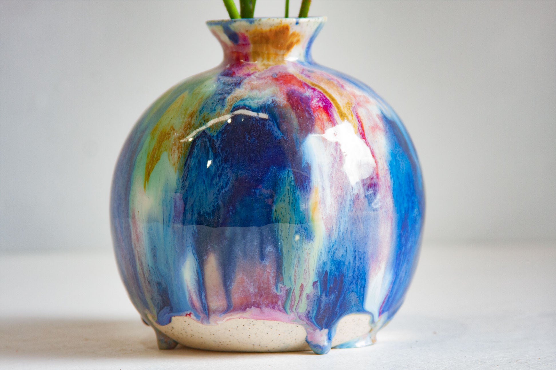 A round ceramic vase with a glossy, dripping multicolor glaze featuring shades of blue, purple, pink, green, and gold, with a small opening at the top containing green stems, set against a plain light gray background.