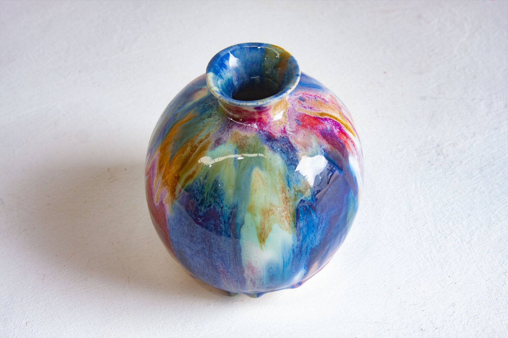 A small round ceramic vase with a glossy finish, featuring a marbled swirl pattern of blue, purple, pink, yellow, and white on a white background.