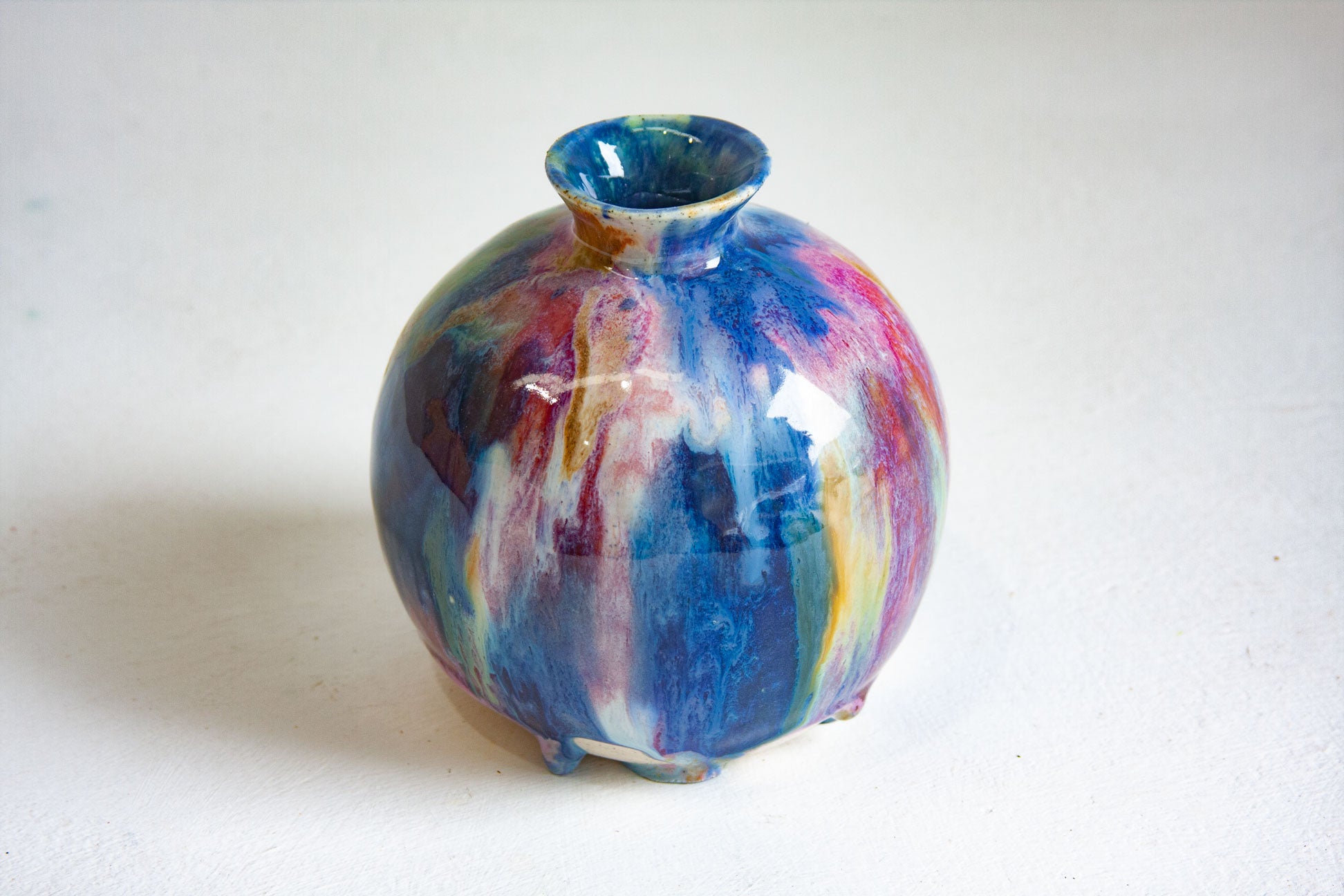 A small round ceramic vase with a flared rim, featuring a glossy marbled glaze with swirls of blue, purple, pink, yellow, and white on a light off-white background.