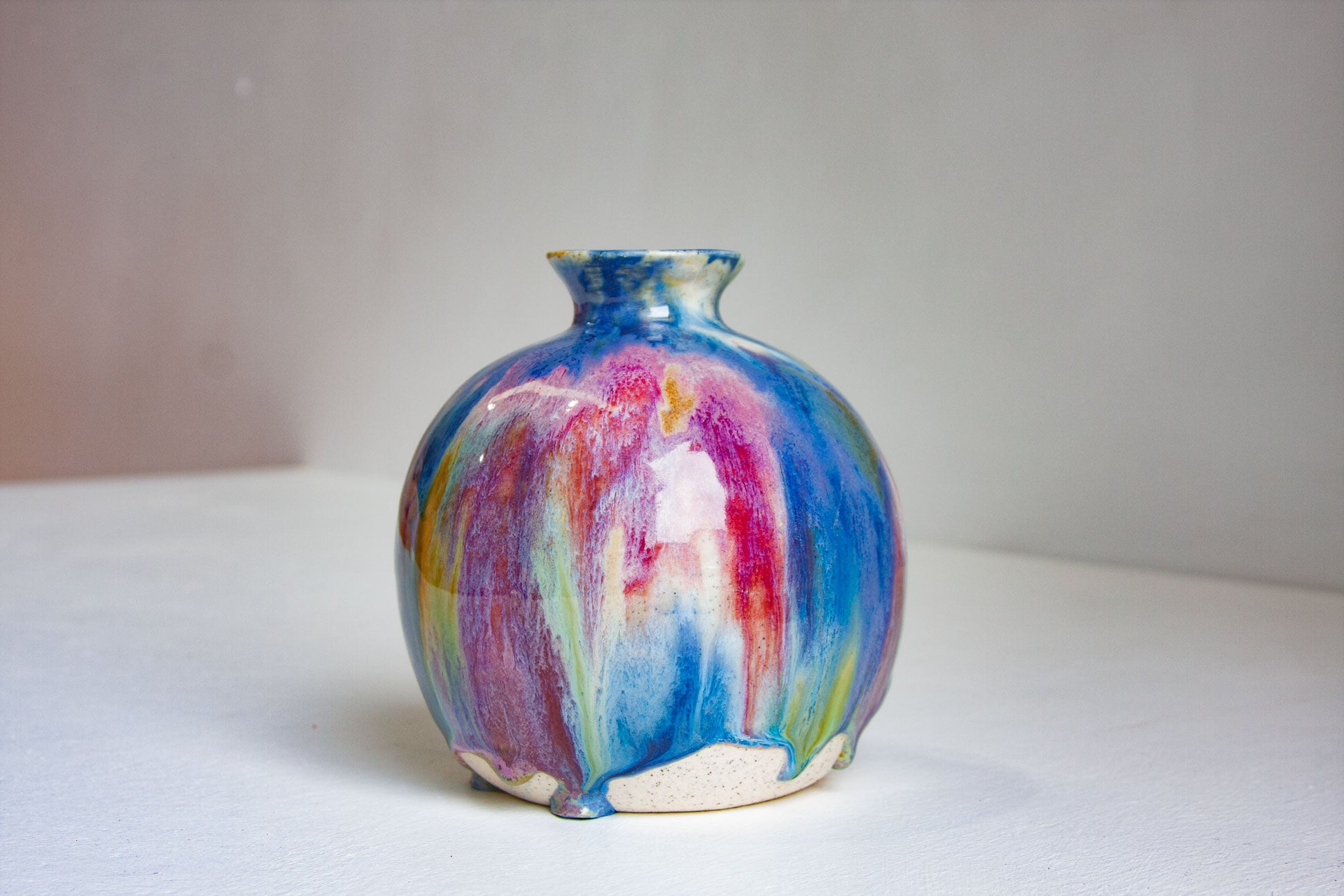 A small ceramic vase with a rounded body and short neck, decorated with a vibrant, multicolored drip glaze featuring blue, purple, pink, green, and yellow on a white surface against a neutral background.