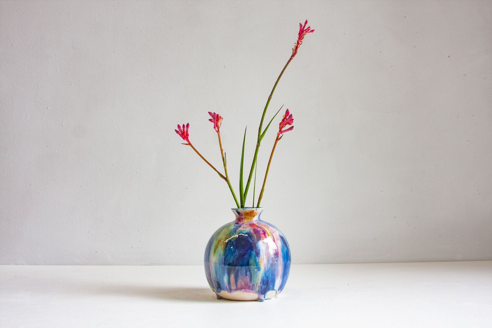 A small round ceramic vase with a colorful marbled blue, purple, and gold pattern, holding two thin green stems with small red flowers, set against a plain light gray background.