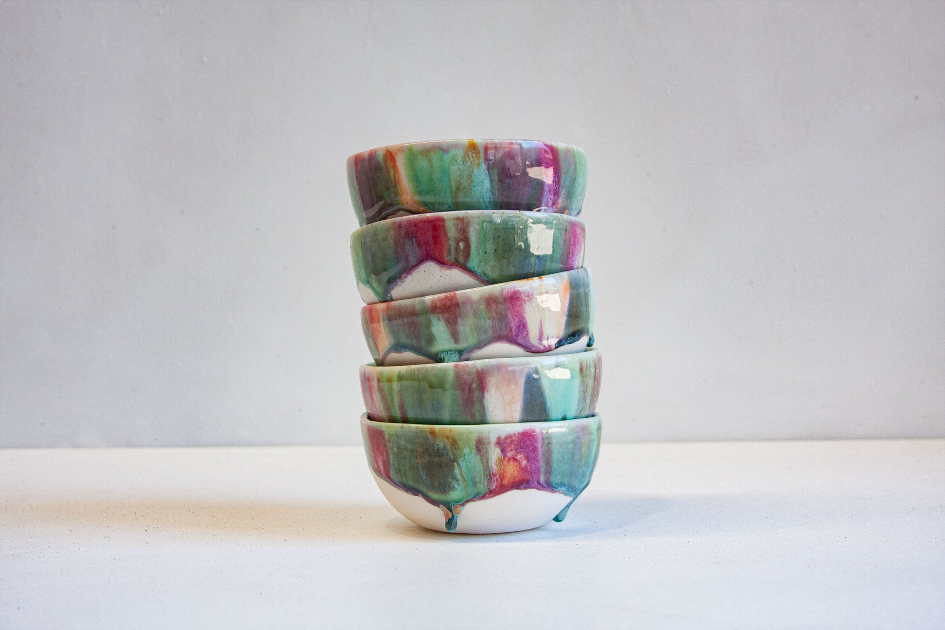 A stack of five small ceramic bowls with a marbled, drip-glazed finish in green, purple, pink, and white, set against a plain light gray background.