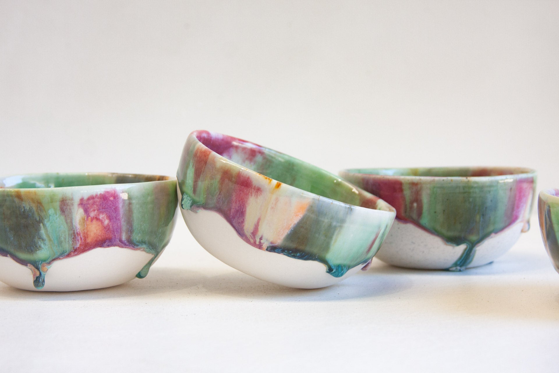 Three small ceramic bowls with a marbled drip glaze pattern in green, purple, pink, and teal on a white base, arranged on a plain white surface against a white background.