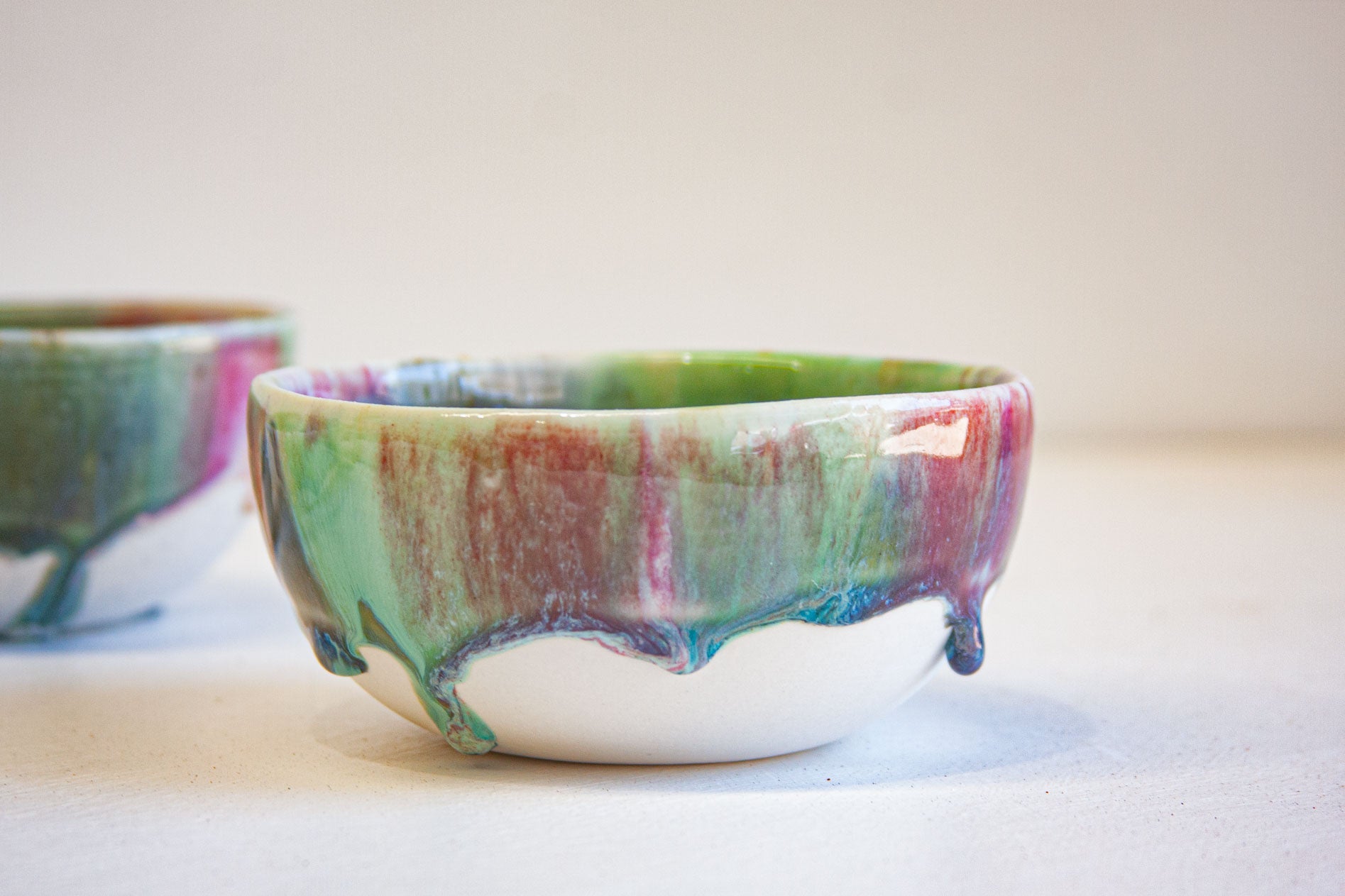 A handmade ceramic bowl with a glossy drip glaze in green, purple, and red over a white base, shown on a neutral light beige background with another blurred bowl in the background.