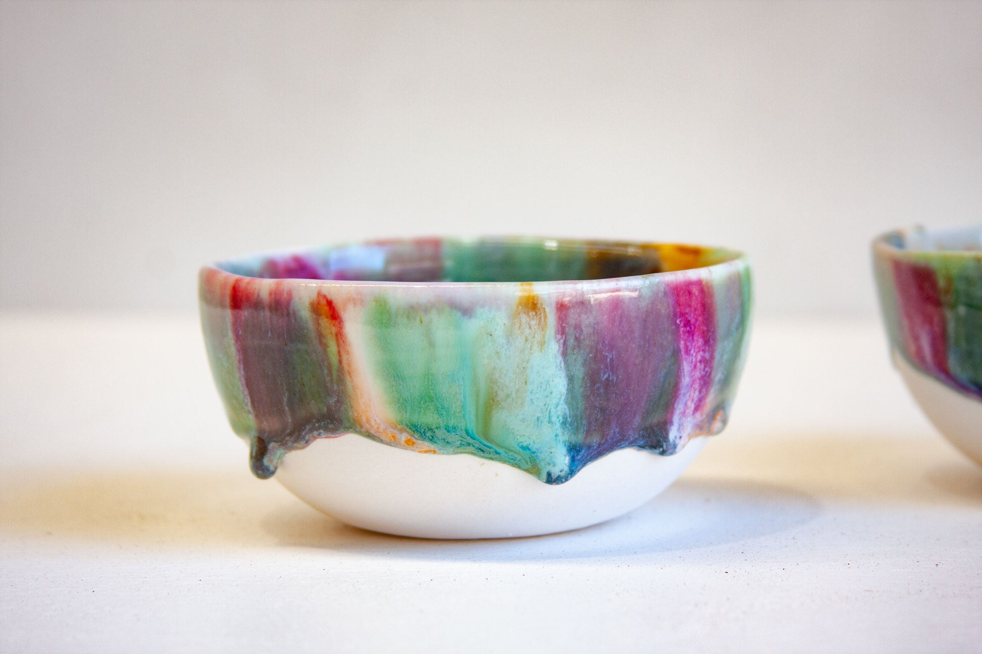 A handmade ceramic bowl with a dripping glaze design, featuring a white base and a rim decorated with colorful paint-like drips of green, teal, purple, pink, and orange, set against a plain light background.