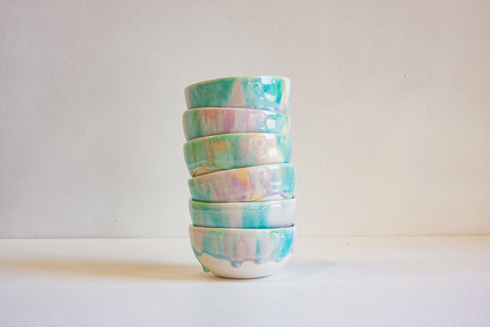 A stack of five small ceramic bowls with a glossy, marbled pastel glaze in shades of teal, mint green, pink, and blue on a white base, set against a plain off-white background.