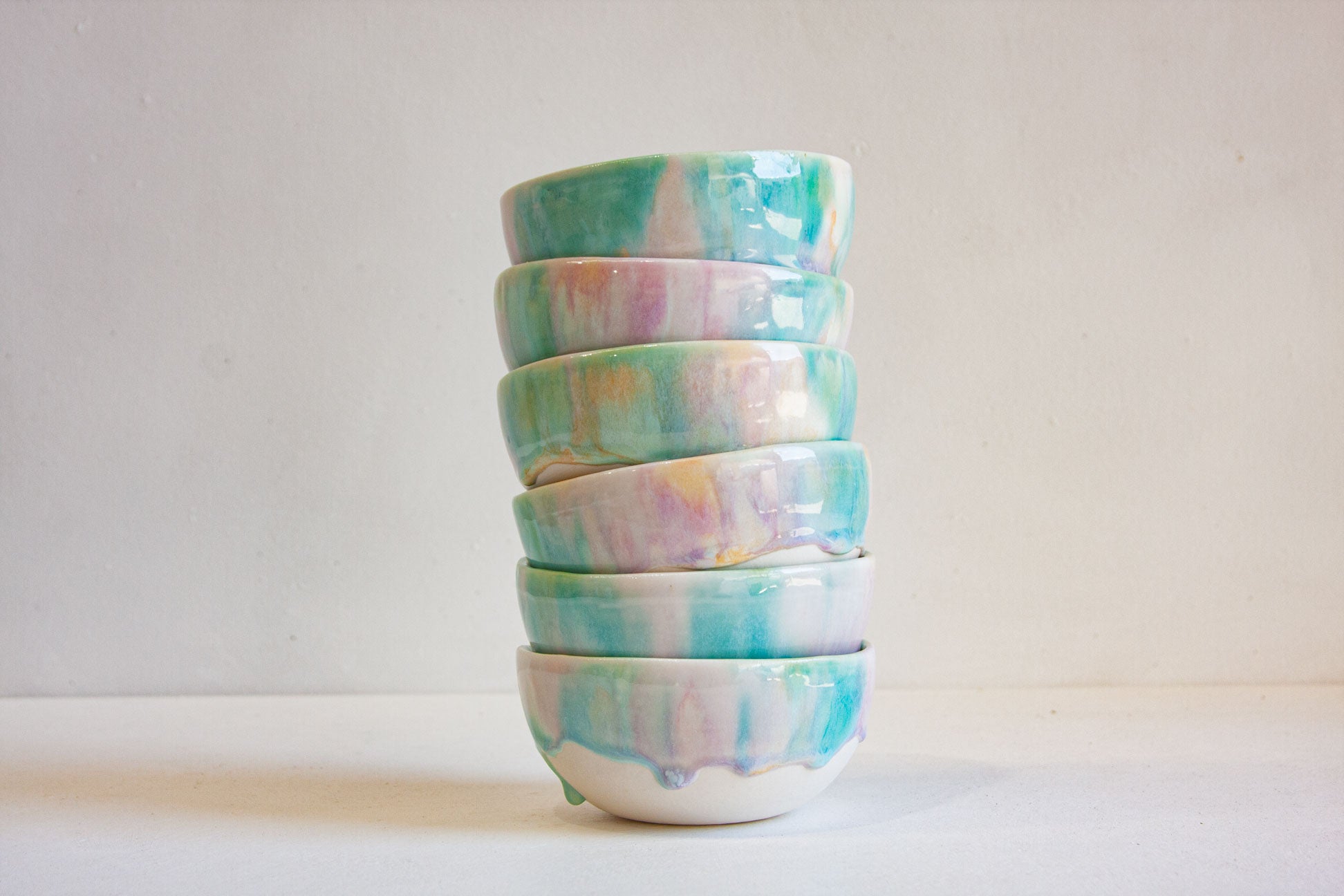 A stack of five small ceramic bowls with a glossy glaze featuring a marbled pattern of turquoise, mint green, lavender, and soft pink on a white base, set against a plain off-white background.