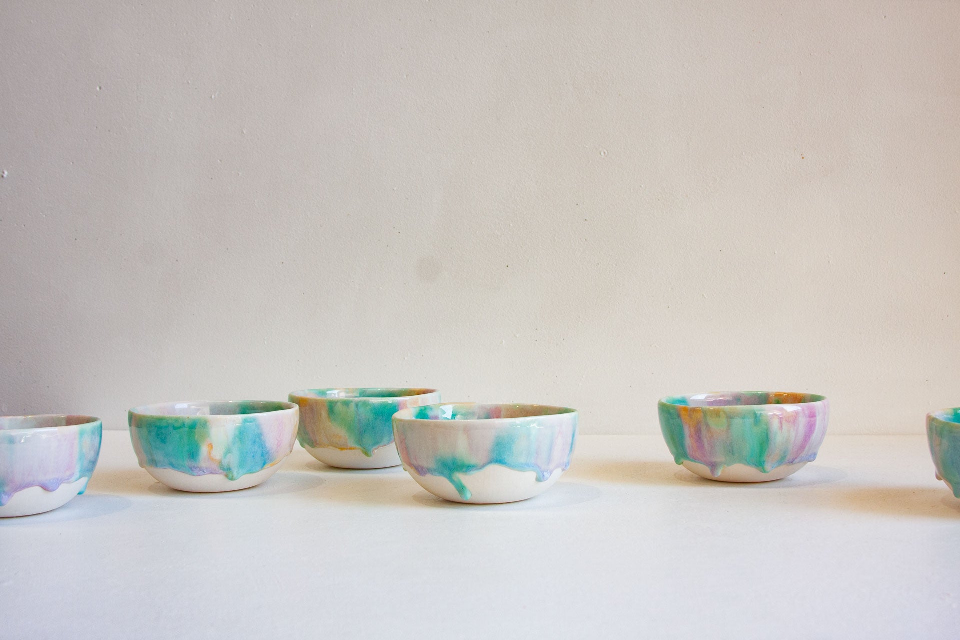 A row of small ceramic bowls with a marbled, watercolor-like pattern in teal, mint green, purple, pink, and orange on an off-white base, set against a plain light beige wall and white surface.