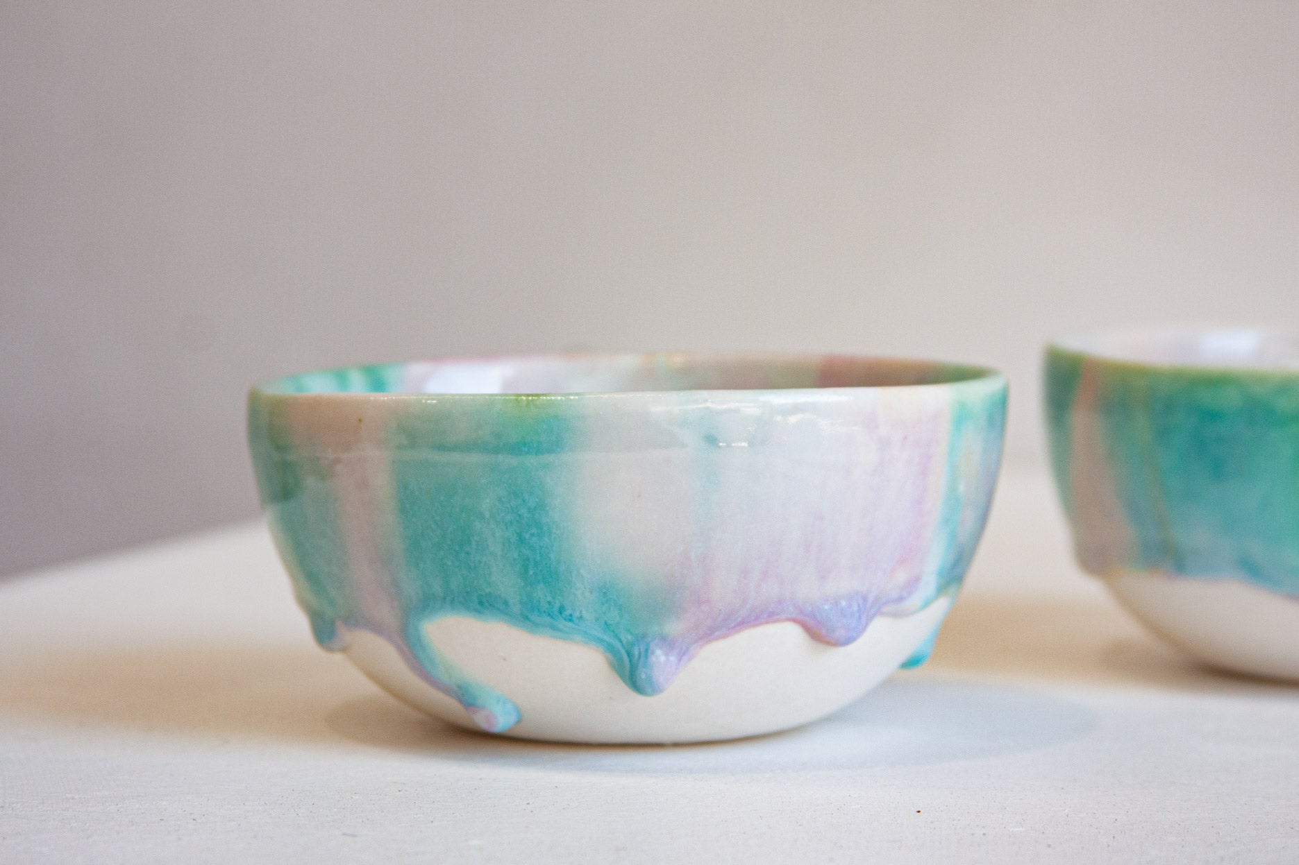 A handmade ceramic bowl with a white base and dripping pastel glaze in shades of teal, green, purple, and pink, shown on a neutral light gray surface with another bowl partially visible in the background.