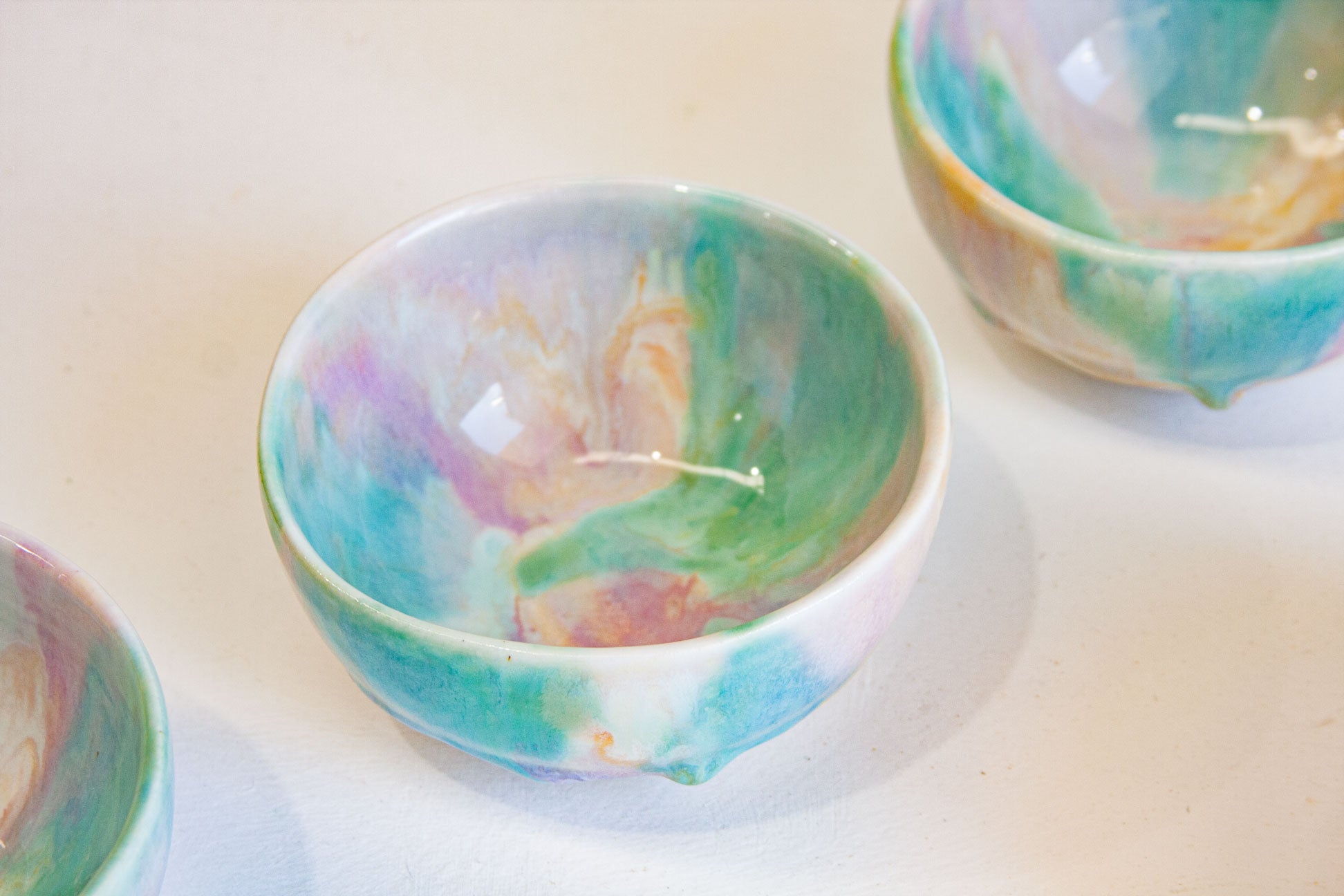 A handmade ceramic bowl with a colorful marbled pattern of teal, green, pink, and purple on a white base, shown on a light surface with other similar bowls partially visible.