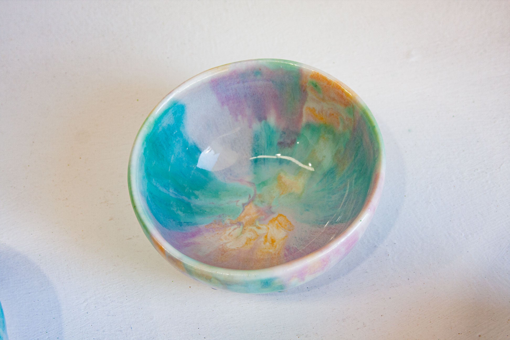 A small handmade ceramic bowl with a colorful marbled pattern in teal, purple, pink, green, and orange, photographed on a light off-white surface.