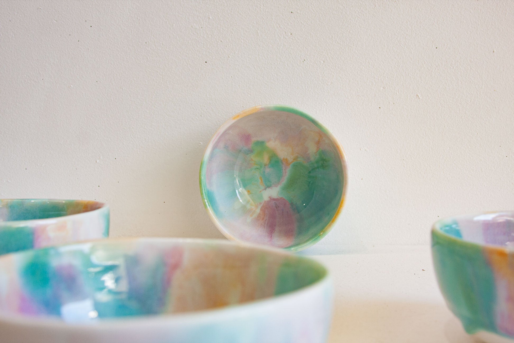 A set of handmade ceramic bowls with a colorful marbled glaze in shades of teal, green, pink, purple, and gold, shown against a plain off-white background.