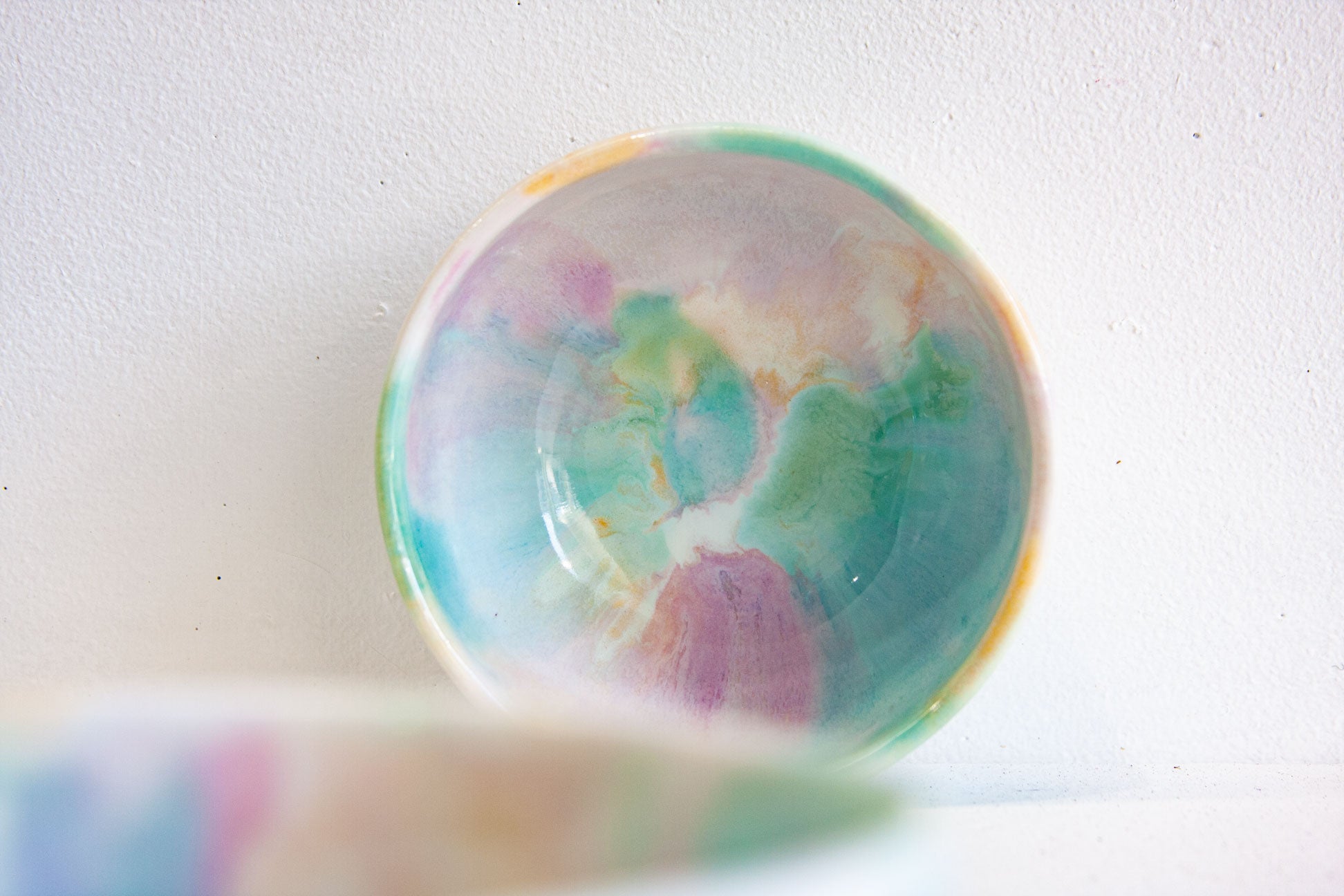 A shallow round bowl with a colorful watercolor-like pattern in shades of teal, green, purple, pink, and yellow on a white background.