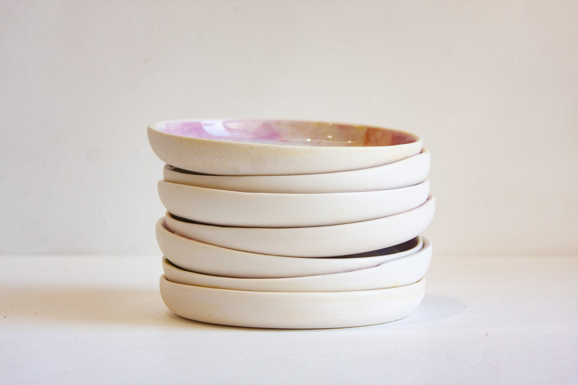 A stack of five shallow ceramic plates with a matte off-white finish and a subtle pastel pink and gold glaze on the top plate, set against a plain light beige background.