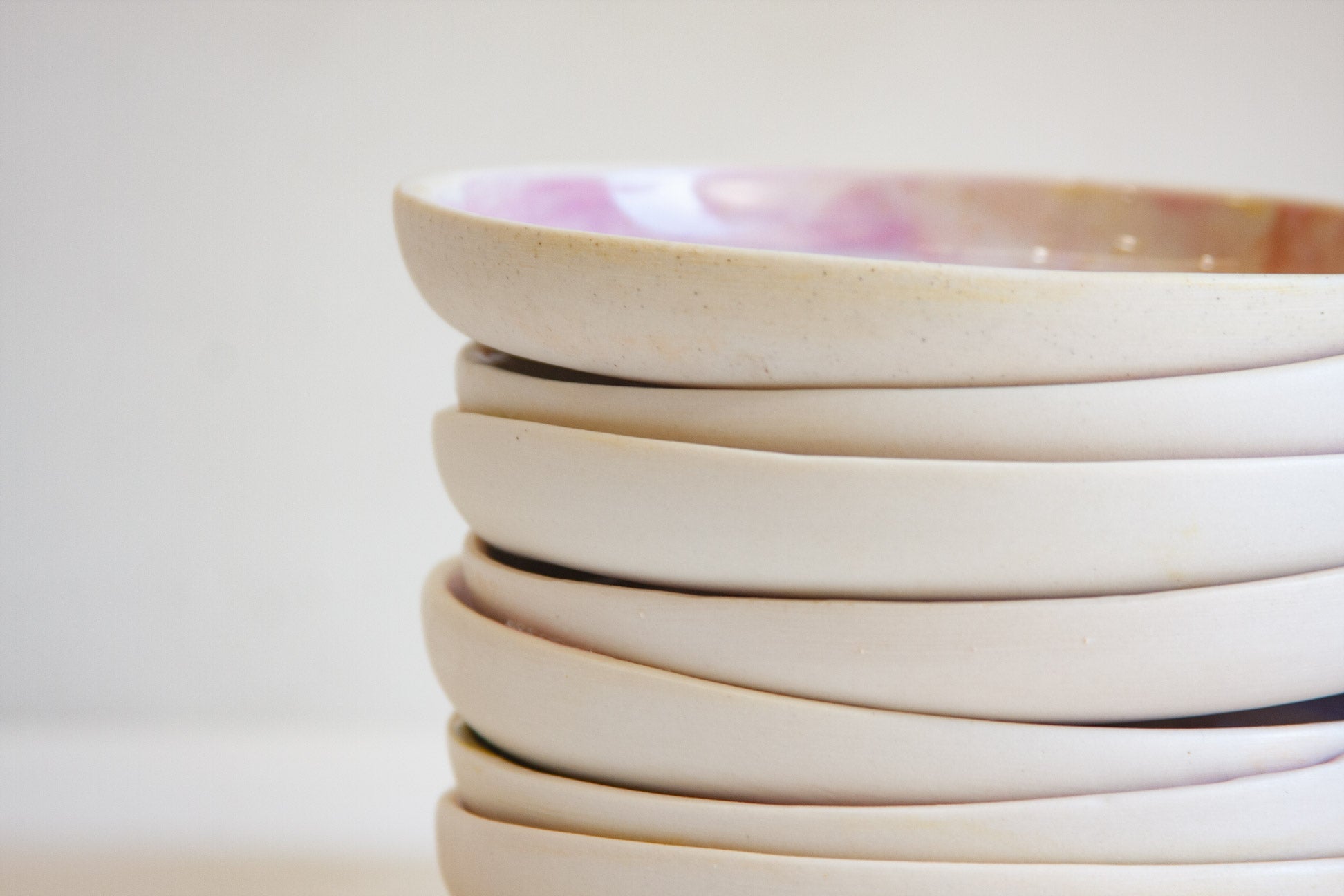 A close-up stack of shallow ceramic plates in a warm off-white or cream glaze, with a top plate showing a soft pink and tan interior glaze, set against a plain light background.