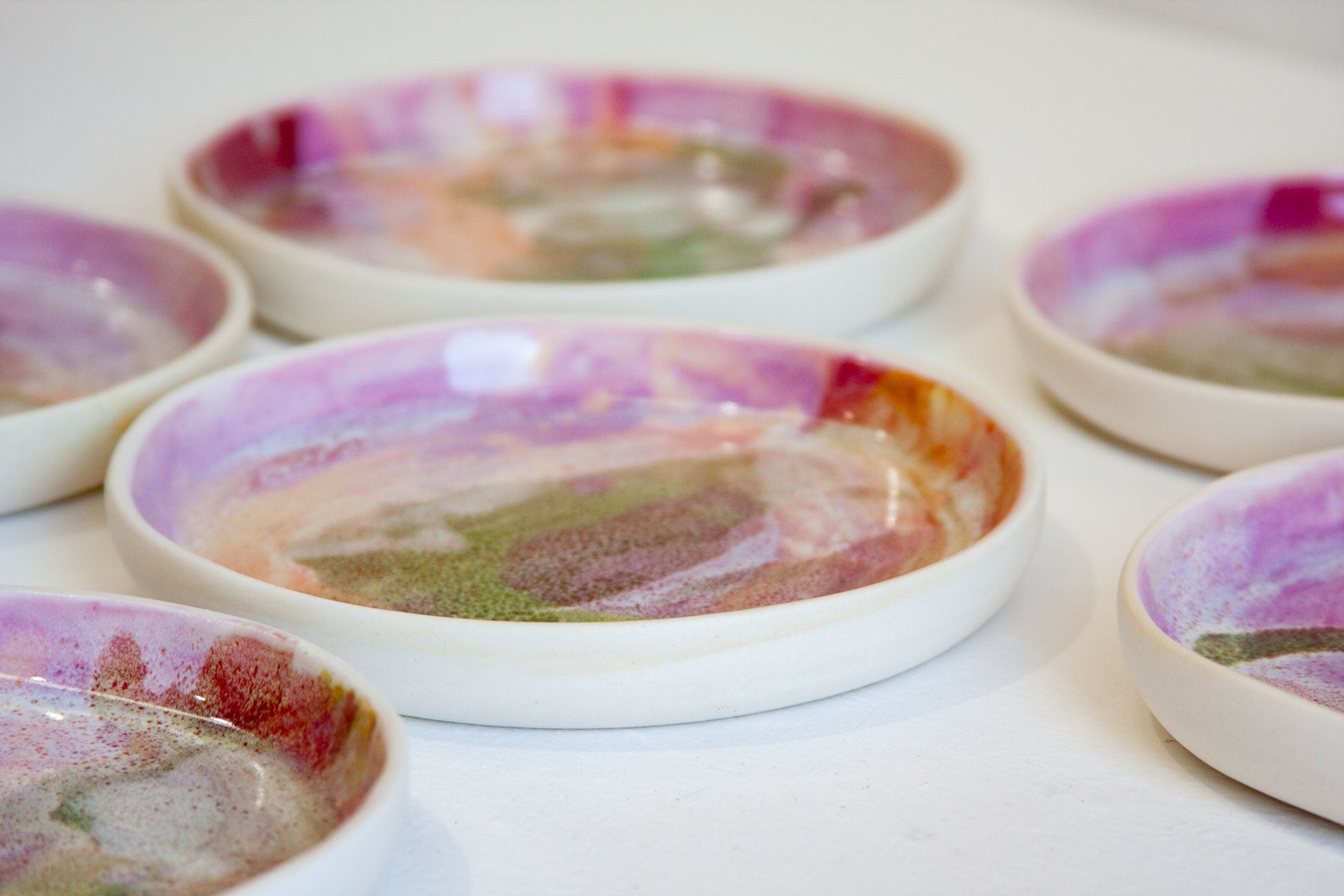 A close-up of several shallow round ceramic dishes with a white base and colorful marbled glaze in pink, purple, green, and brown tones, arranged on a white surface.