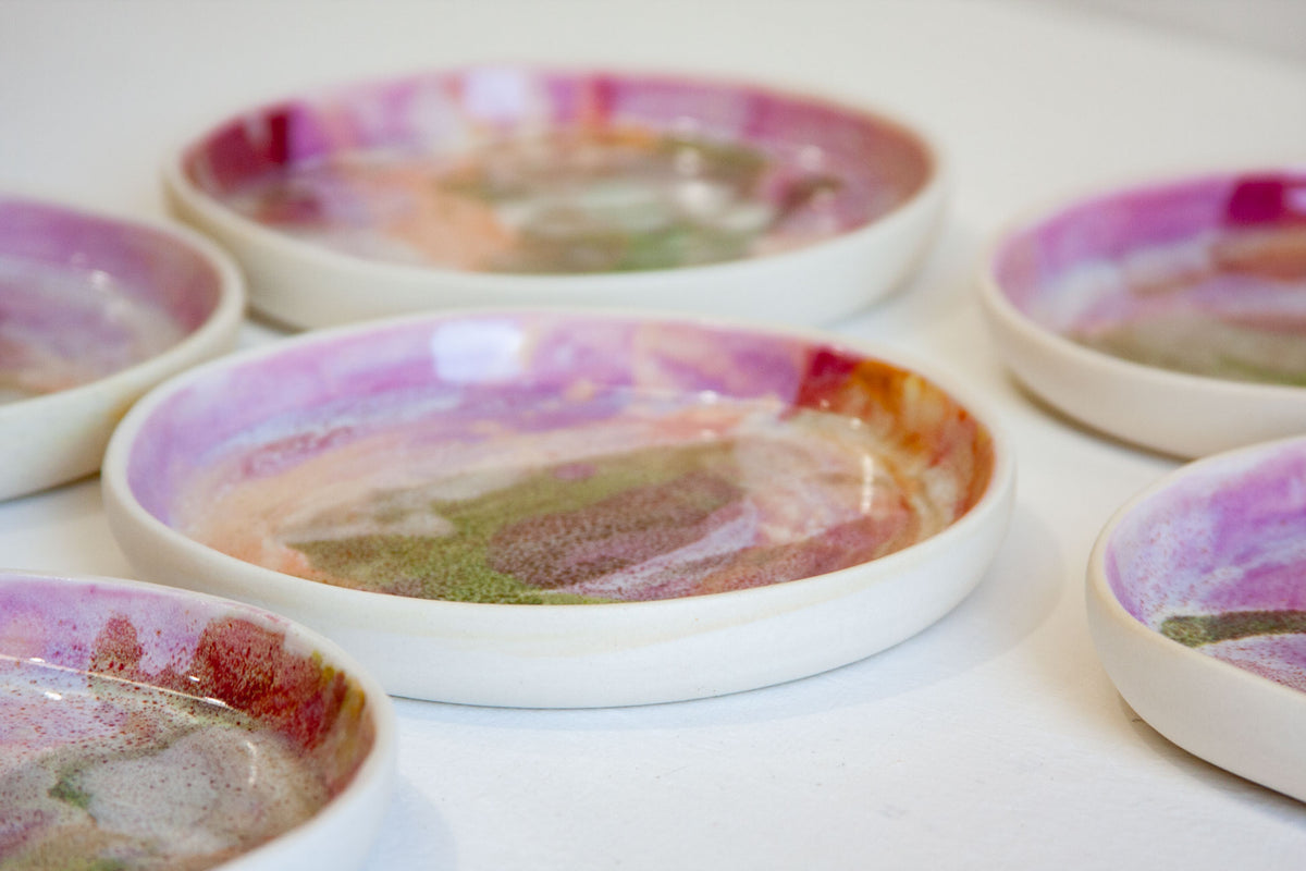 A close-up of several shallow round ceramic dishes with a white base and colourful marbled glaze in pink, purple, green, and brown tones, arranged on a white surface.