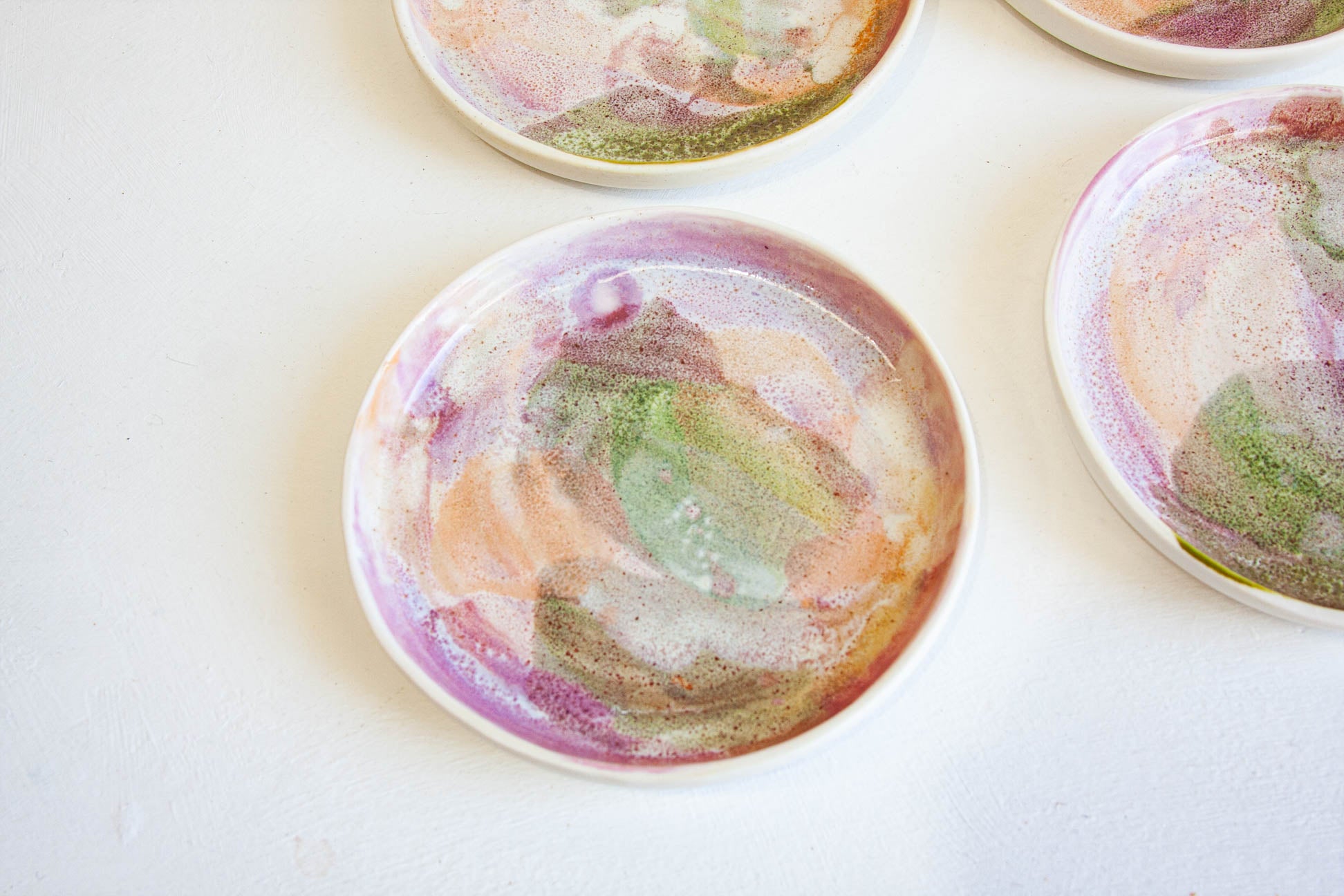 Three round ceramic plates with colorful abstract painted designs in pink, purple, green, and orange on a white background.
