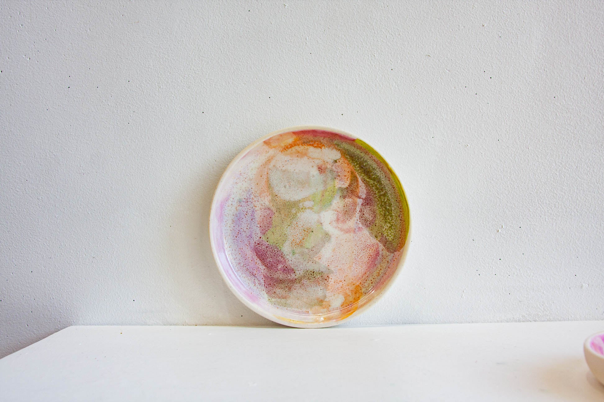 A handmade ceramic plate with a colorful abstract painted design in pink, orange, yellow, and green, resting on a white surface against a textured white wall.