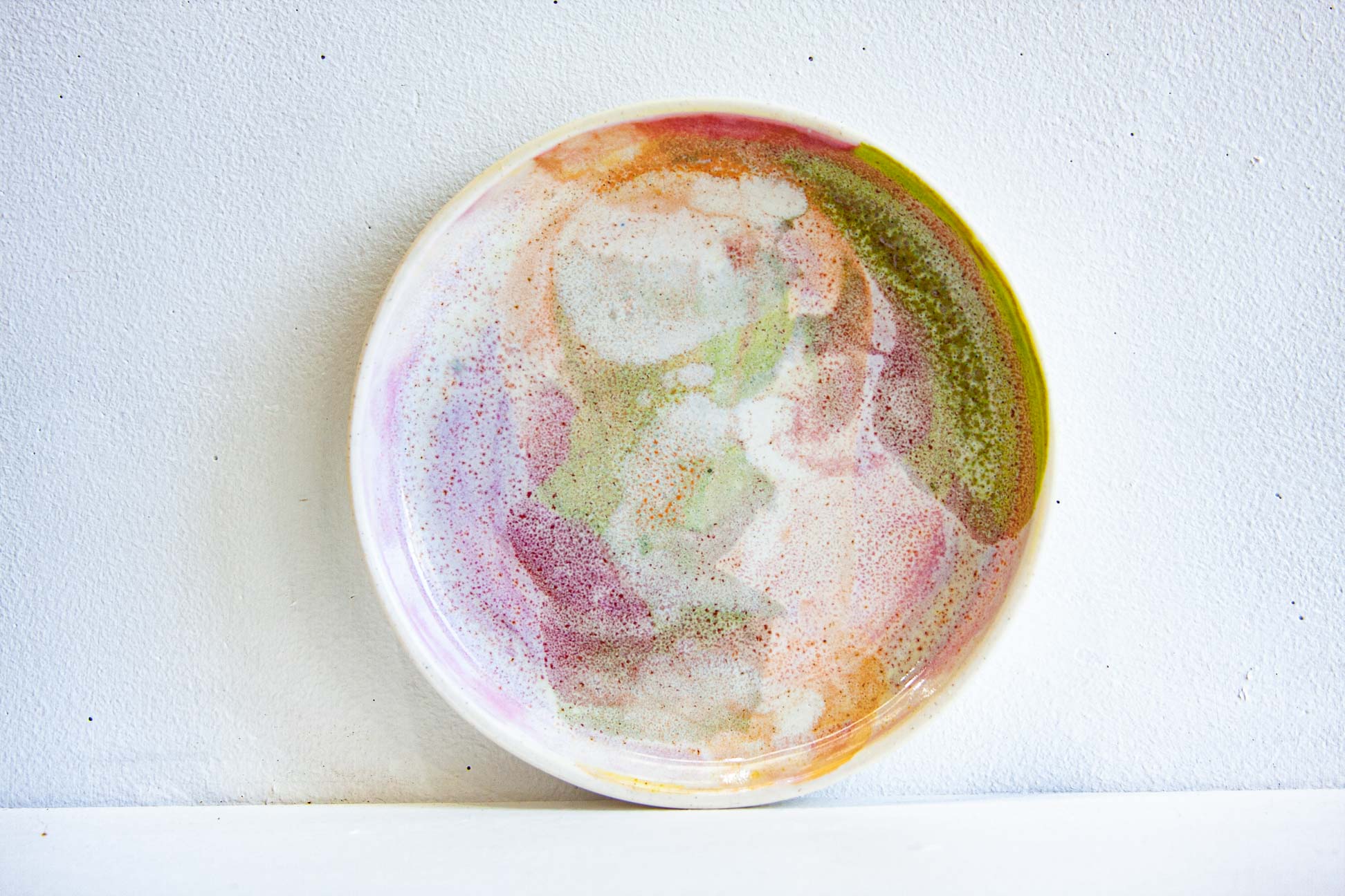A handmade ceramic plate with a colorful, abstract hand-painted design in shades of pink, green, orange, and purple on a white background.