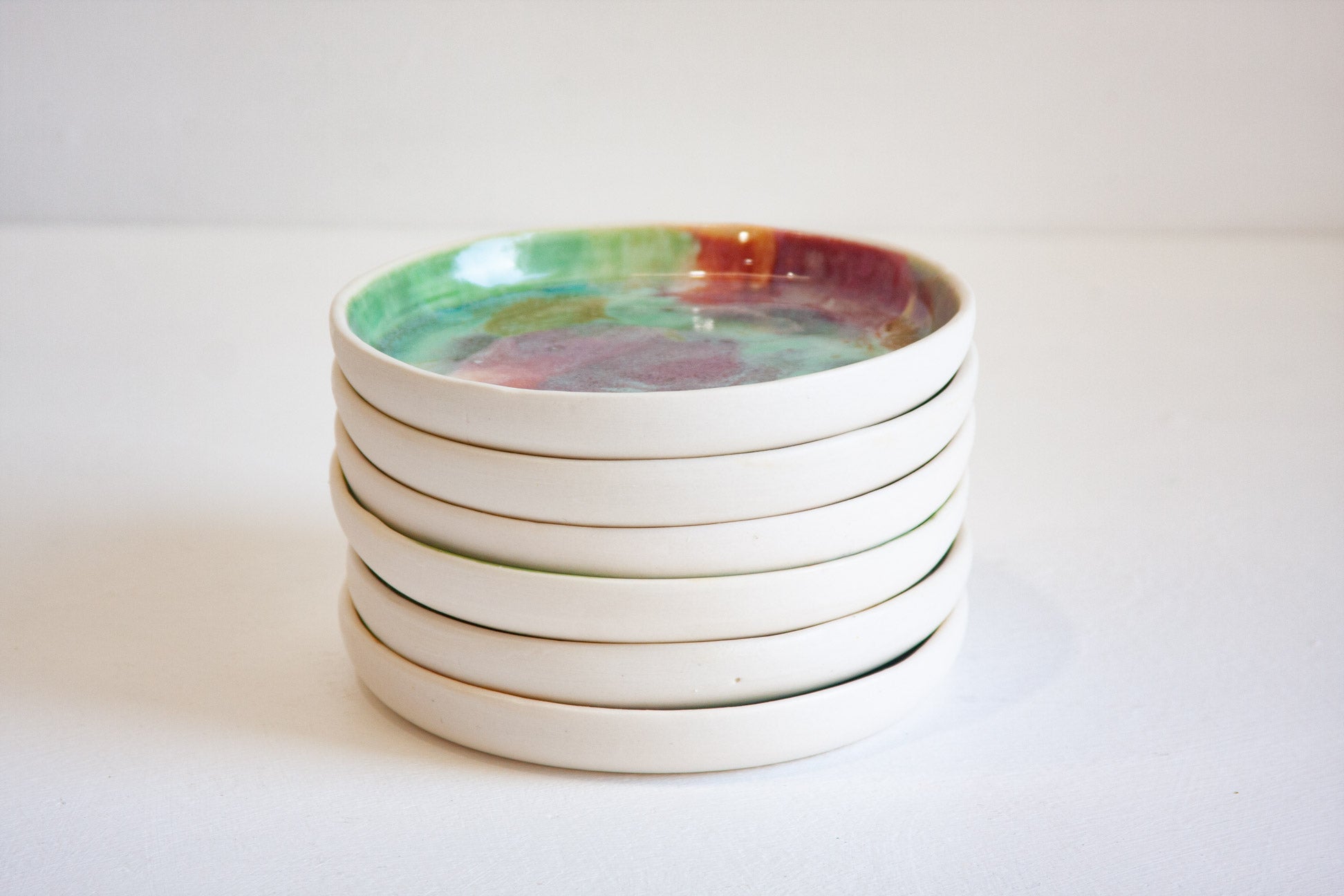 A stack of six round ceramic plates with a glossy, hand-painted multicolour glaze on the top coaster, set against a plain white background.