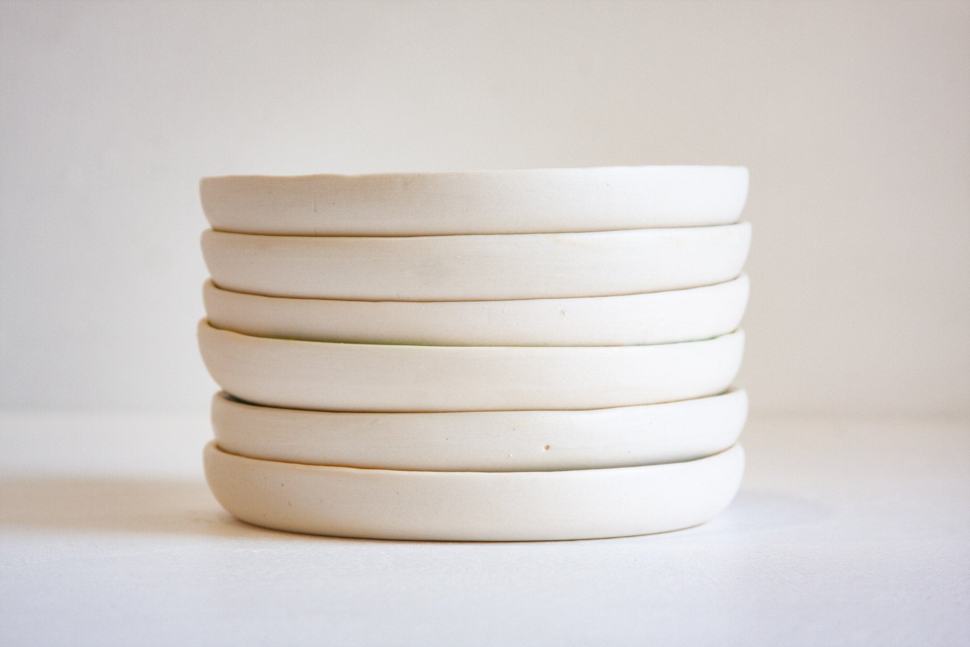 A stack of six handmade ceramic plates in a warm off-white, matte finish, photographed against a neutral light beige background.