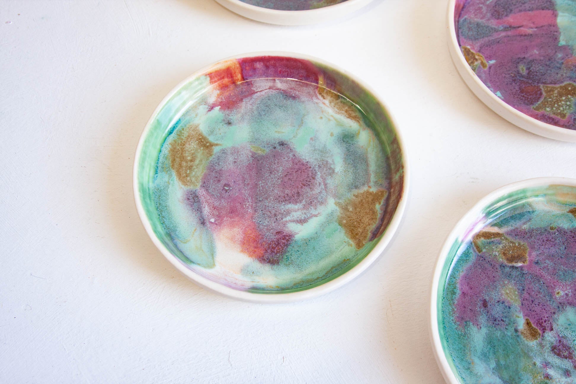 A close-up of a round ceramic plate with a colourful marbled pattern in purple, teal, pink, and gold on a white surface, with other partially visible plates around it.