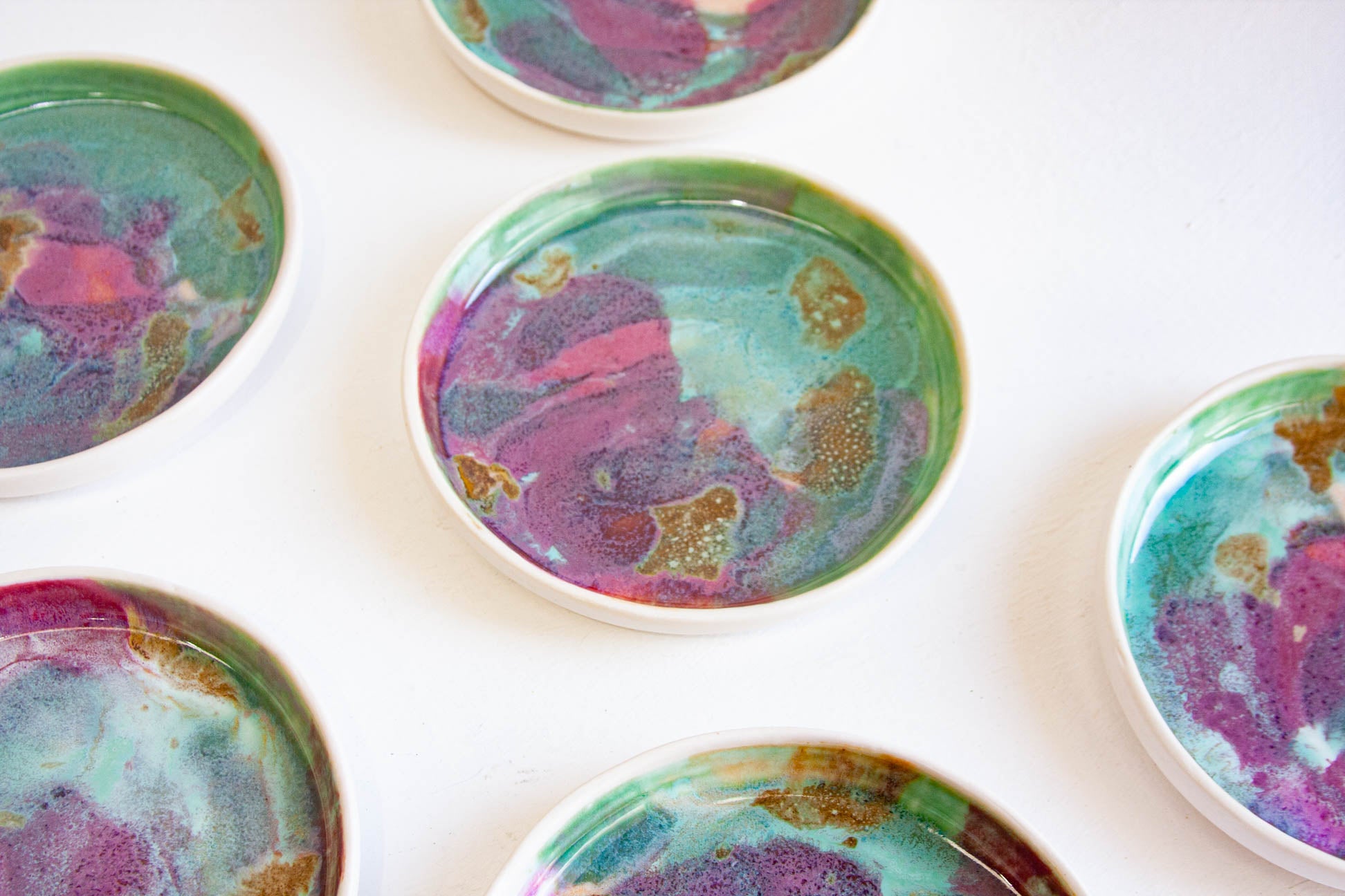 A close-up overhead view of several round shallow dishes or bowls with colourful marbled tops in teal, purple, pink, and gold, arranged on a white surface.