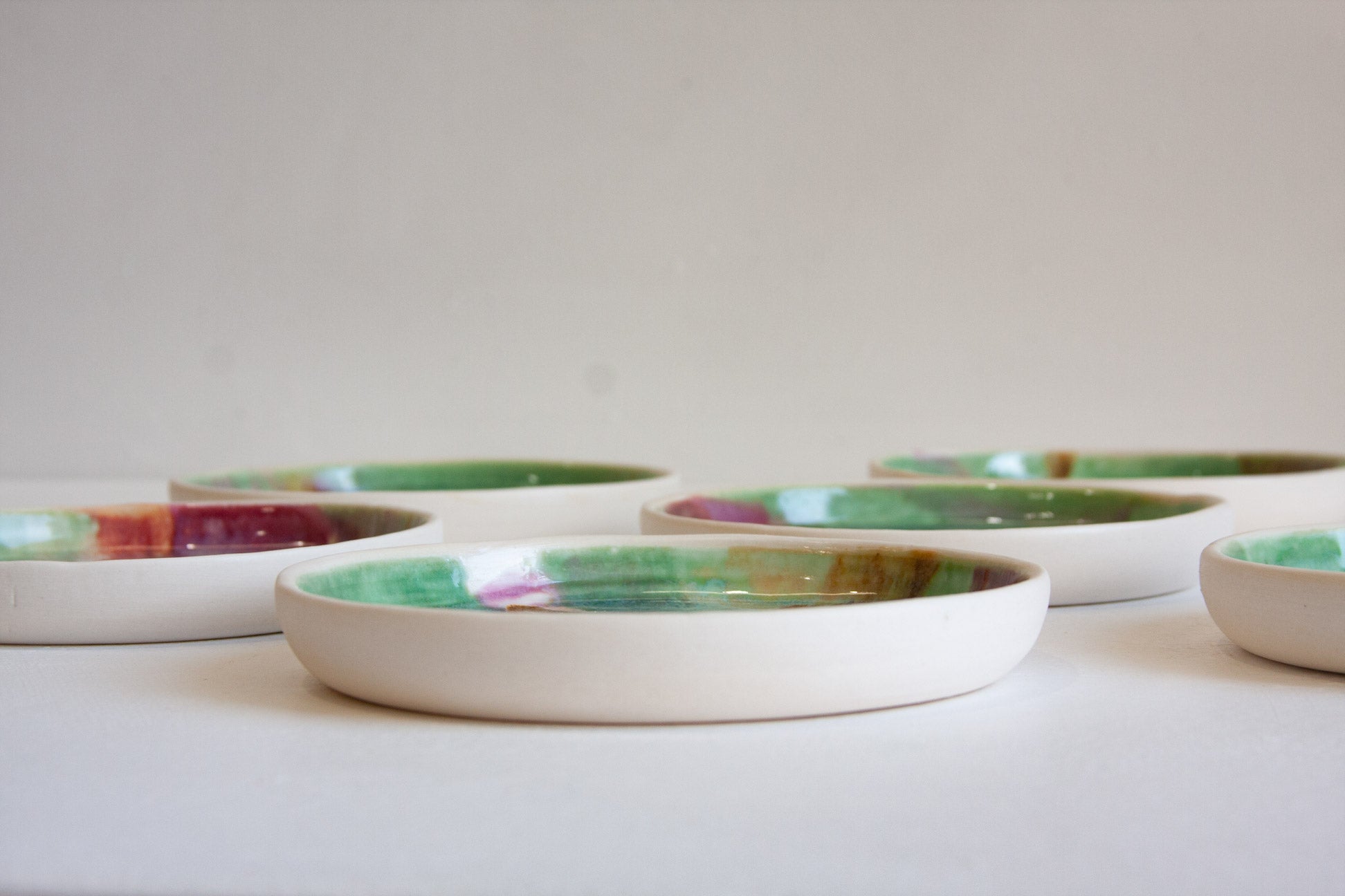 A set of shallow round ceramic dishes with a matte off-white exterior and a glossy, marbled interior featuring green, purple, and gold tones, arranged on a light surface against a neutral background.
