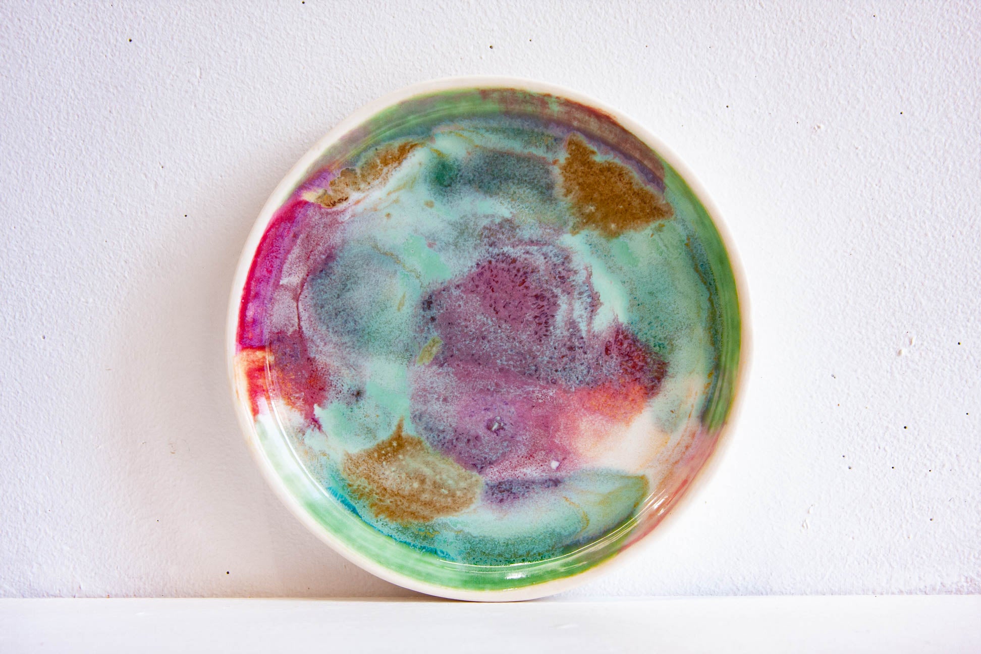 A round ceramic plate with a colourful abstract watercolour design in shades of purple, green, teal, and gold on a white background.