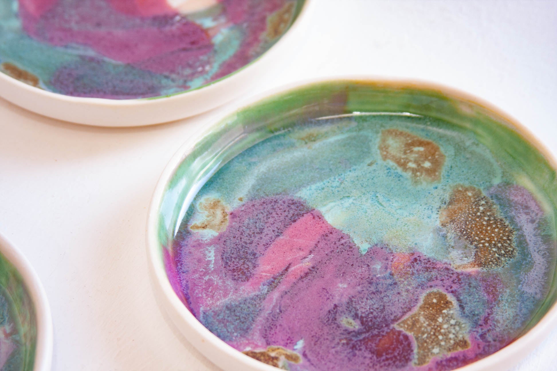 A close-up of a shallow round bowl with a colourful marbled glaze in purple, teal, green, and brown on a white surface, with partial views of similar bowls at the top and left edges.