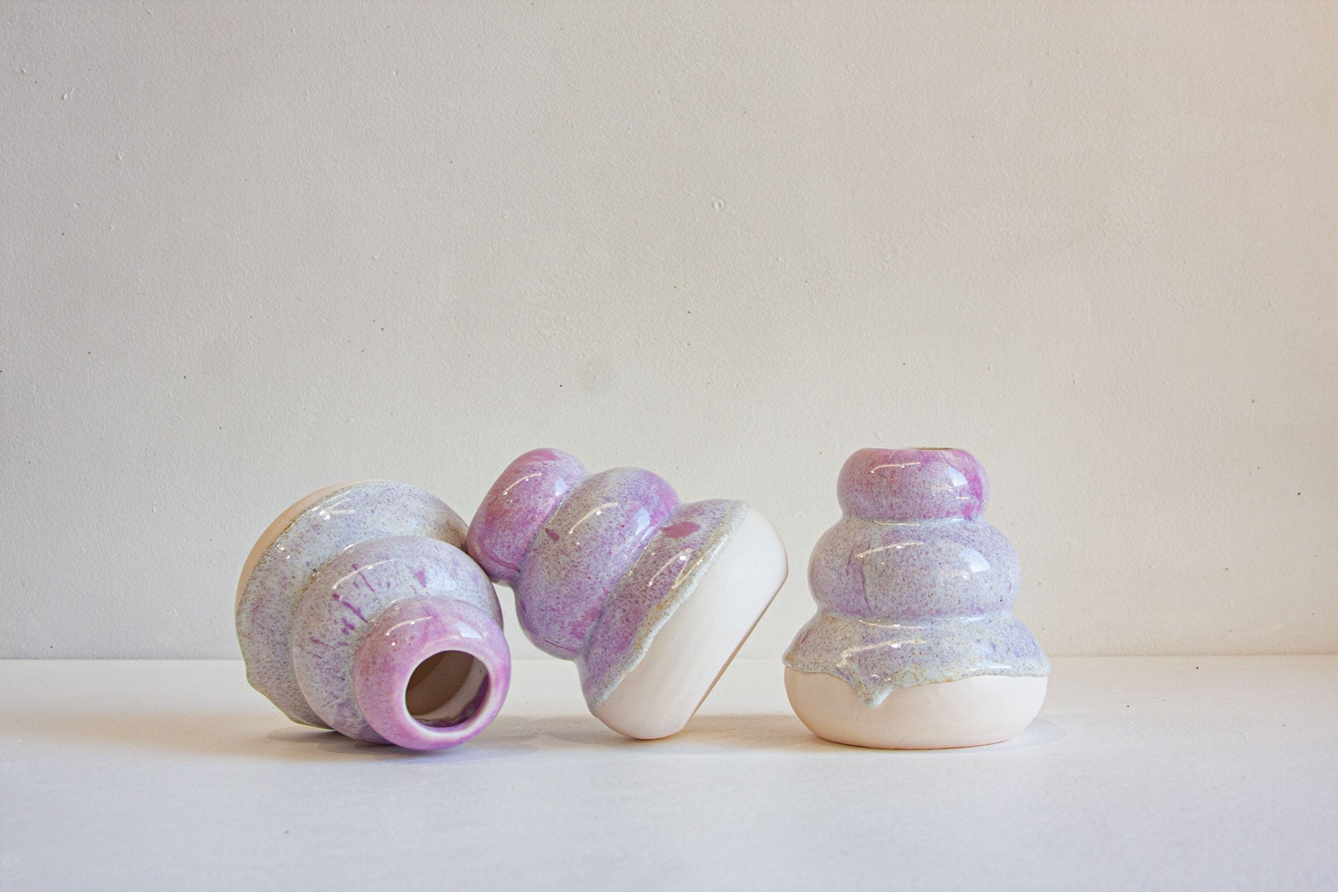Three small decorative ceramic vases with a pastel lavender, purple, and white iridescent glaze, arranged on a white surface against a light beige wall.