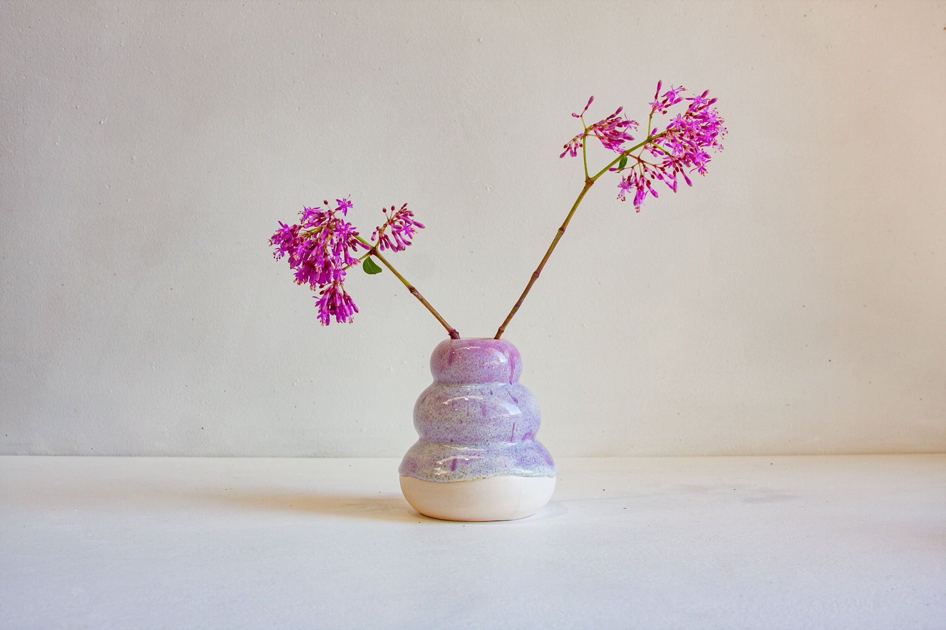A small handmade ceramic bud vase with a lavender and white marbled glaze, holding two stems of bright pink flowers, set against a neutral cream background.