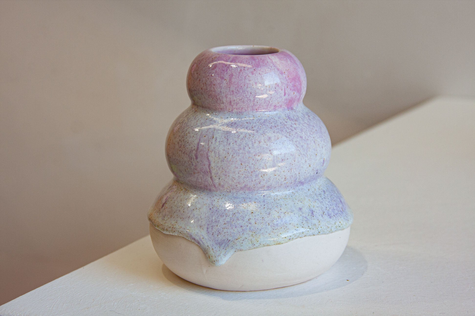 A small ceramic bud vase with three stacked rounded tiers, featuring a glossy pastel glaze that transitions from lavender and pink at the top to pale blue and white at the base, shown on a neutral tabletop.