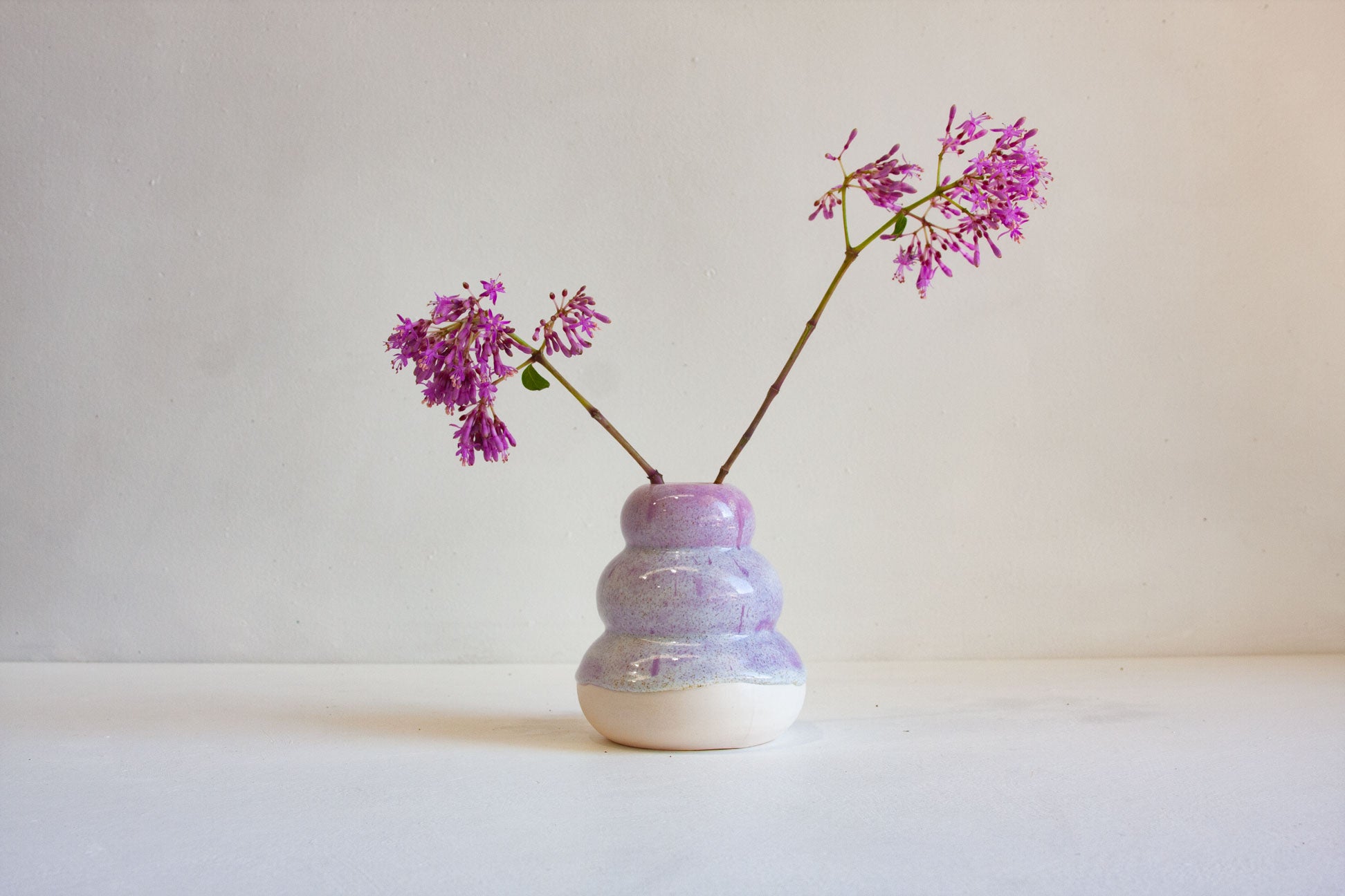 A small ceramic vase with a stacked, rounded design in lavender, purple, and white glaze, holding two thin stems with small purple flowers, set against a plain off-white background.