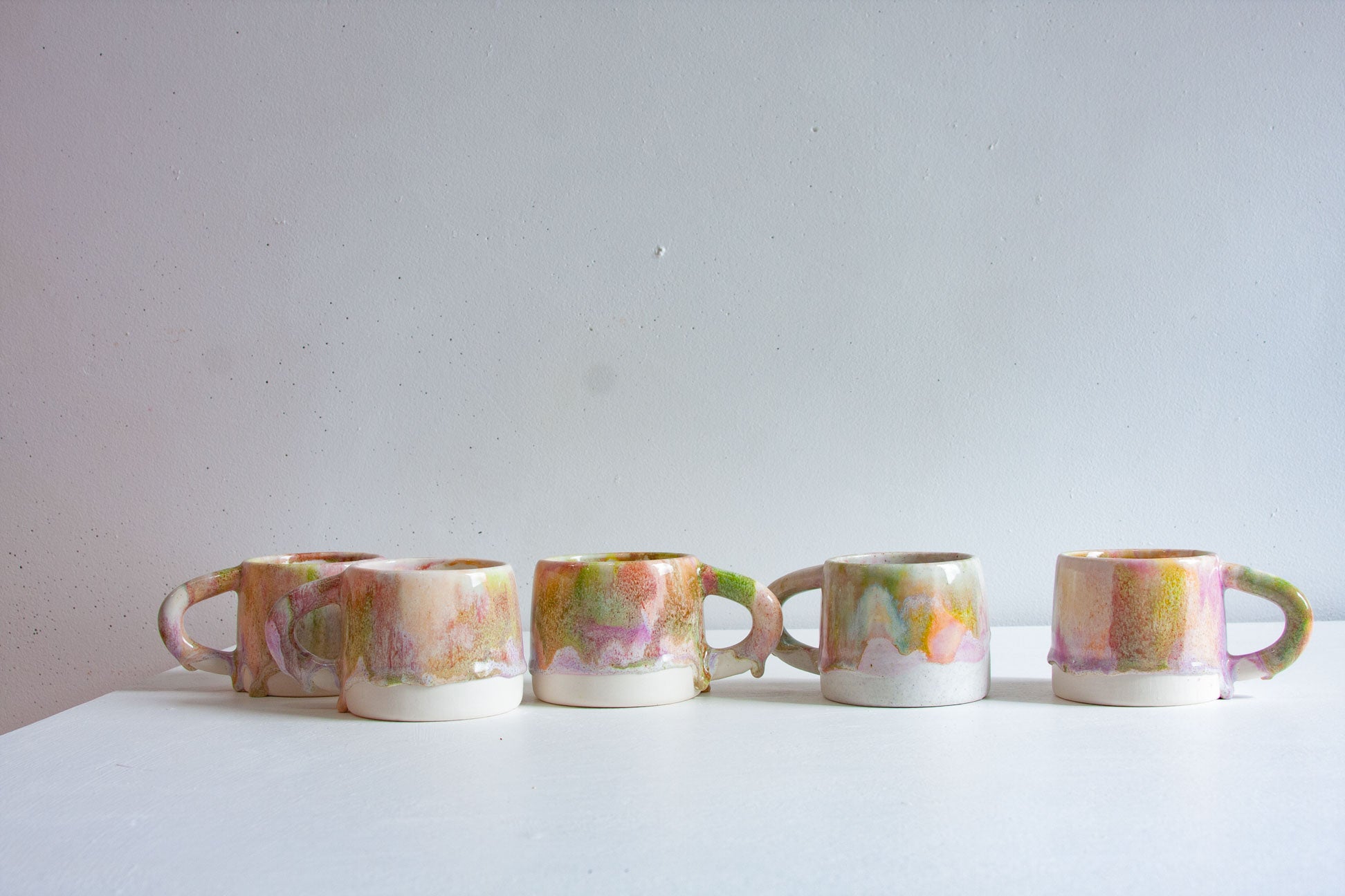 Five handmade ceramic mugs in a row, brush-glazed in mottled peach, pink, green and lilac above bare white clay bases.