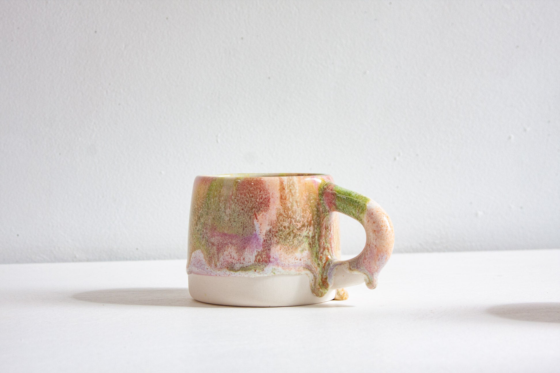 A single handmade ceramic mug with a chunky handle, glazed in mottled peach, green and lilac above a bare white clay base.