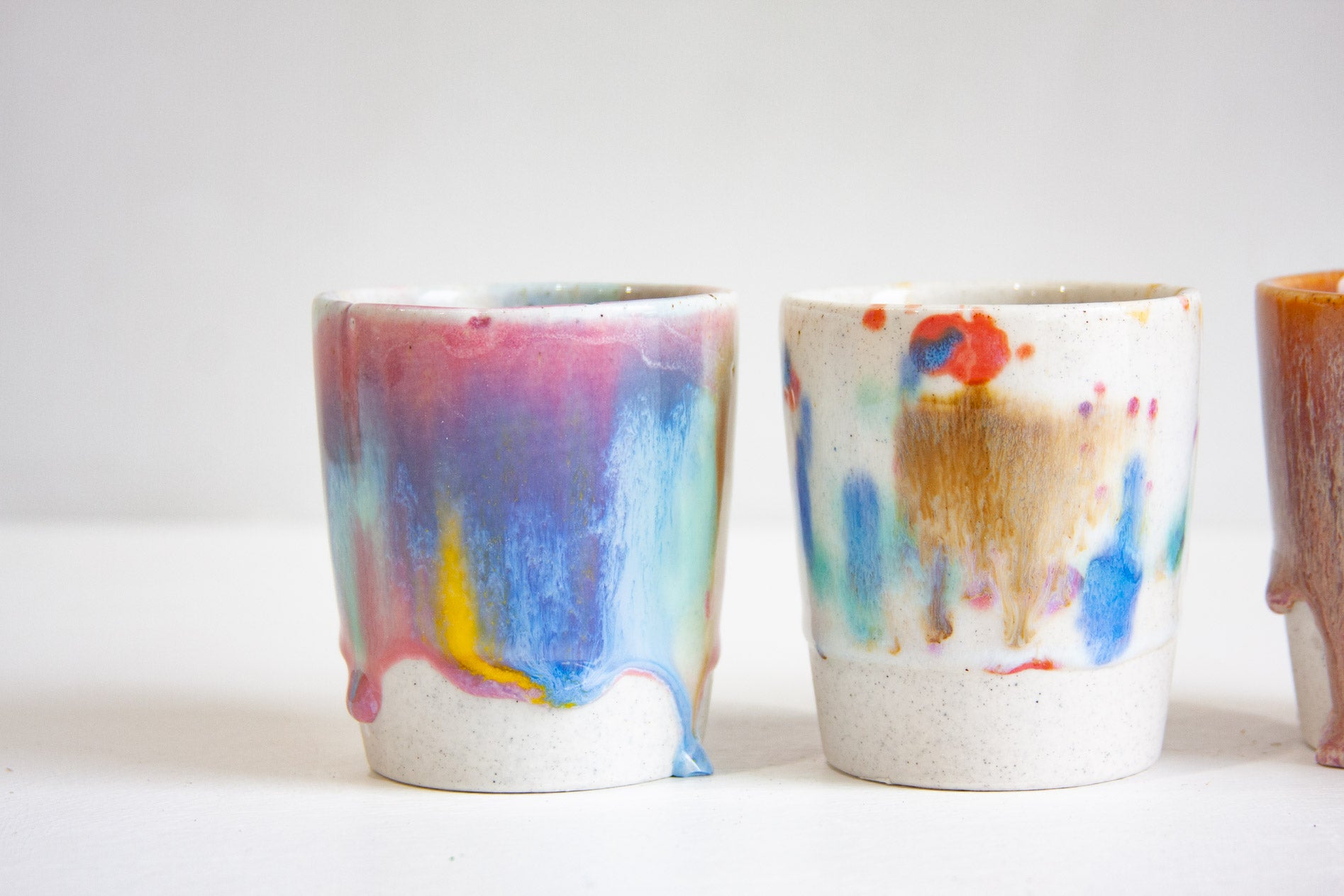 Two handmade ceramic tumblers close up, one glazed in drips of pink, blue, teal and yellow, the other speckled white flecked with blue, red and ochre, both on unglazed speckled bases.
