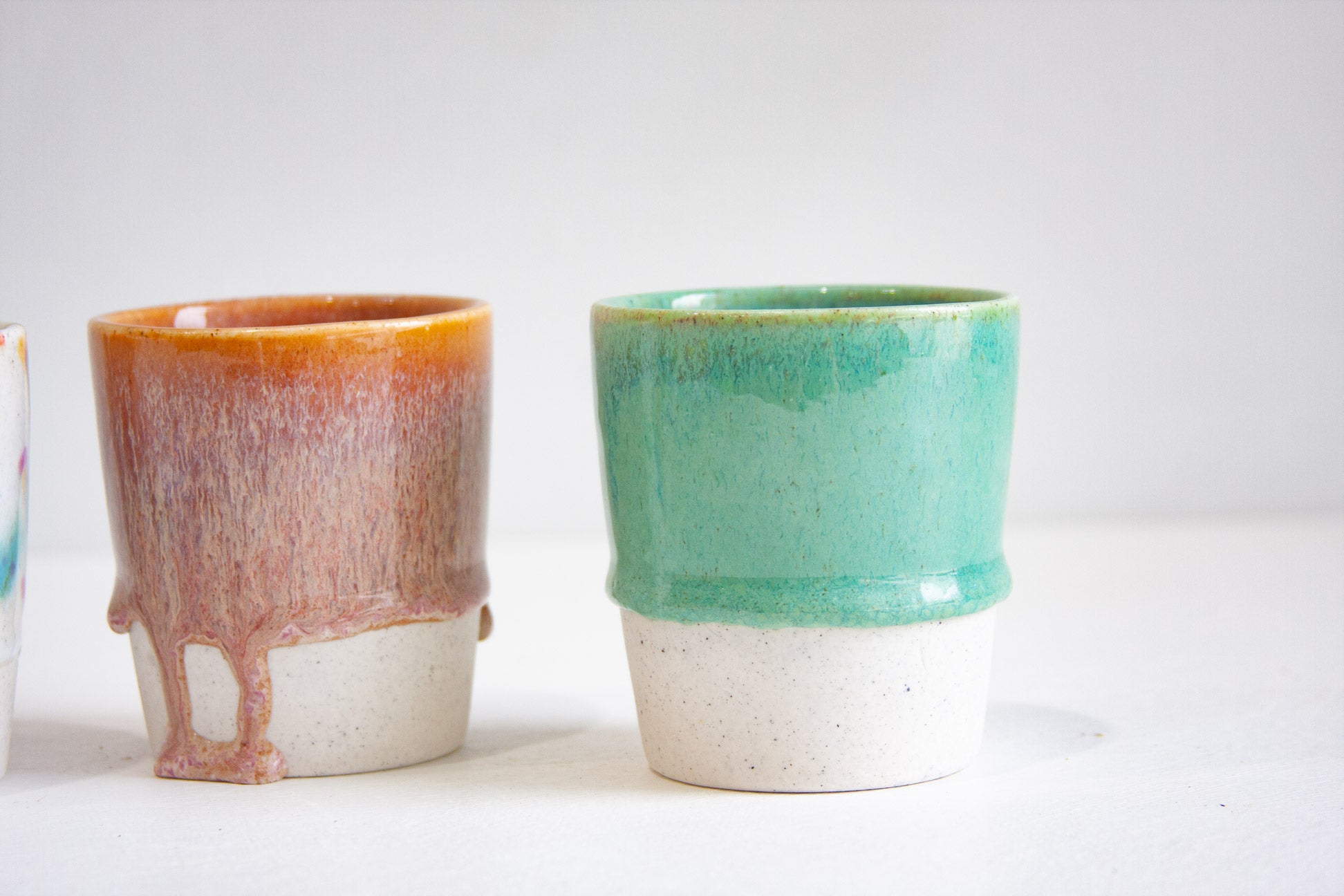 Two handmade ceramic tumblers close up, one in a rust orange glaze running down into mauve, the other in solid jade green, both on unglazed speckled bases.