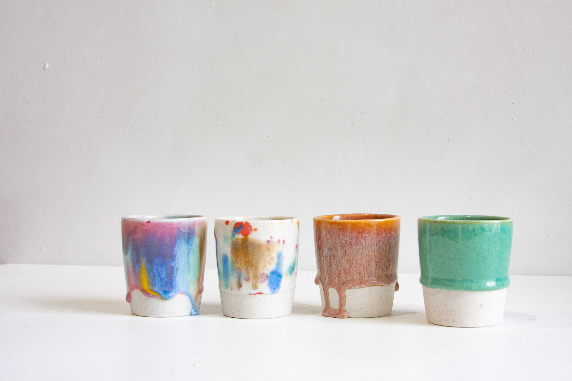 Four handmade ceramic tumblers in a row, one in a drippy multicolour glaze, one speckled white with flecks of blue and red, one rust orange and one jade green, each on an unglazed speckled base.