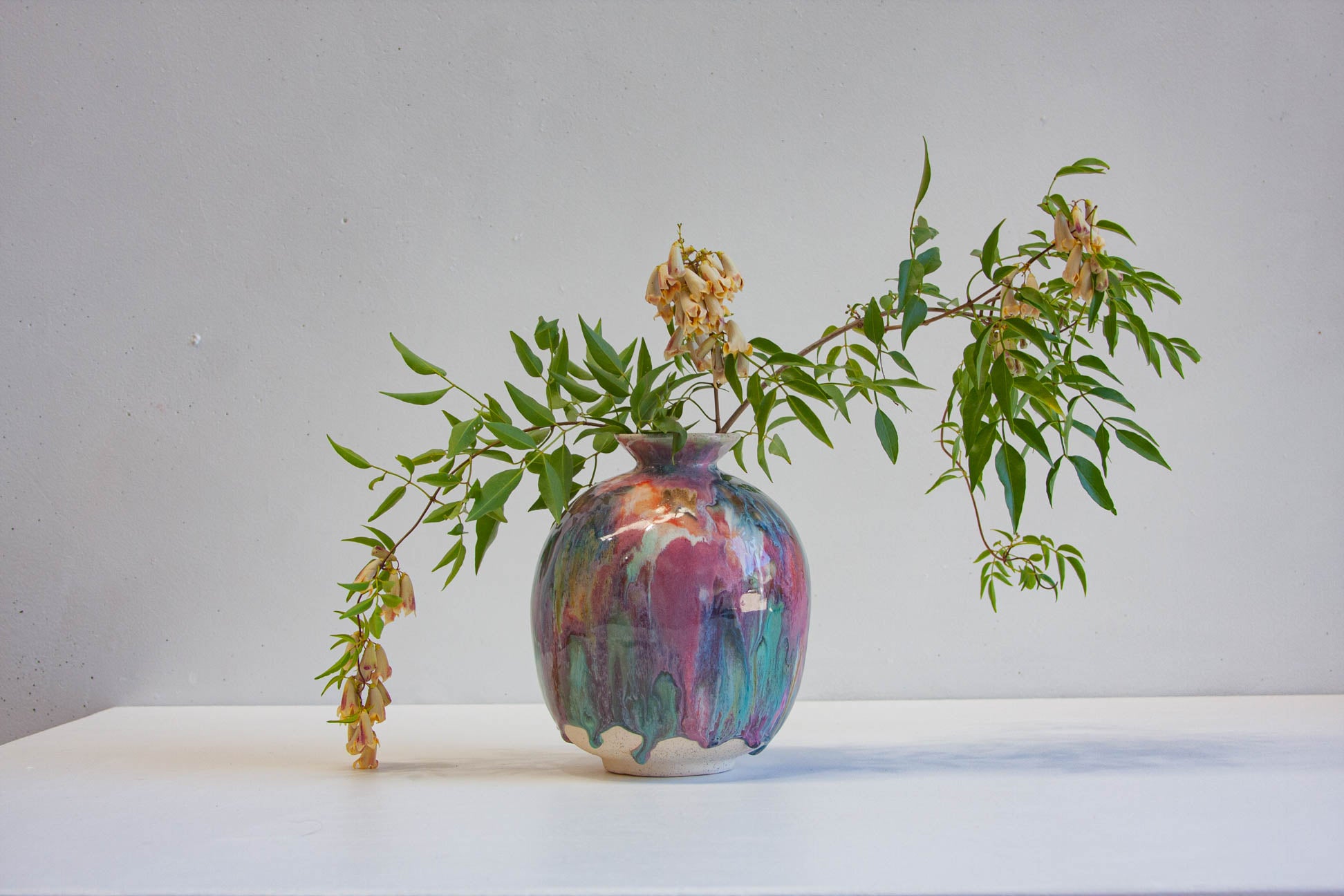 A ceramic vase with a marbled, dripping glaze in purple, teal, and cream, holding green leafy branches against a plain light gray wall and white surface.