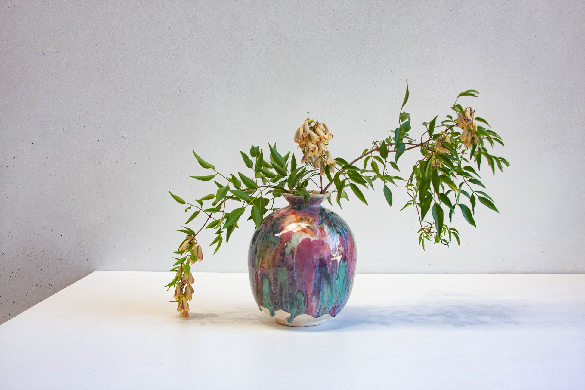 A small ceramic vase with a rounded body and narrow flared neck, decorated with a glossy multicolour drip glaze in pink, purple, blue, green, orange, and red tones, photographed against a plain light background.