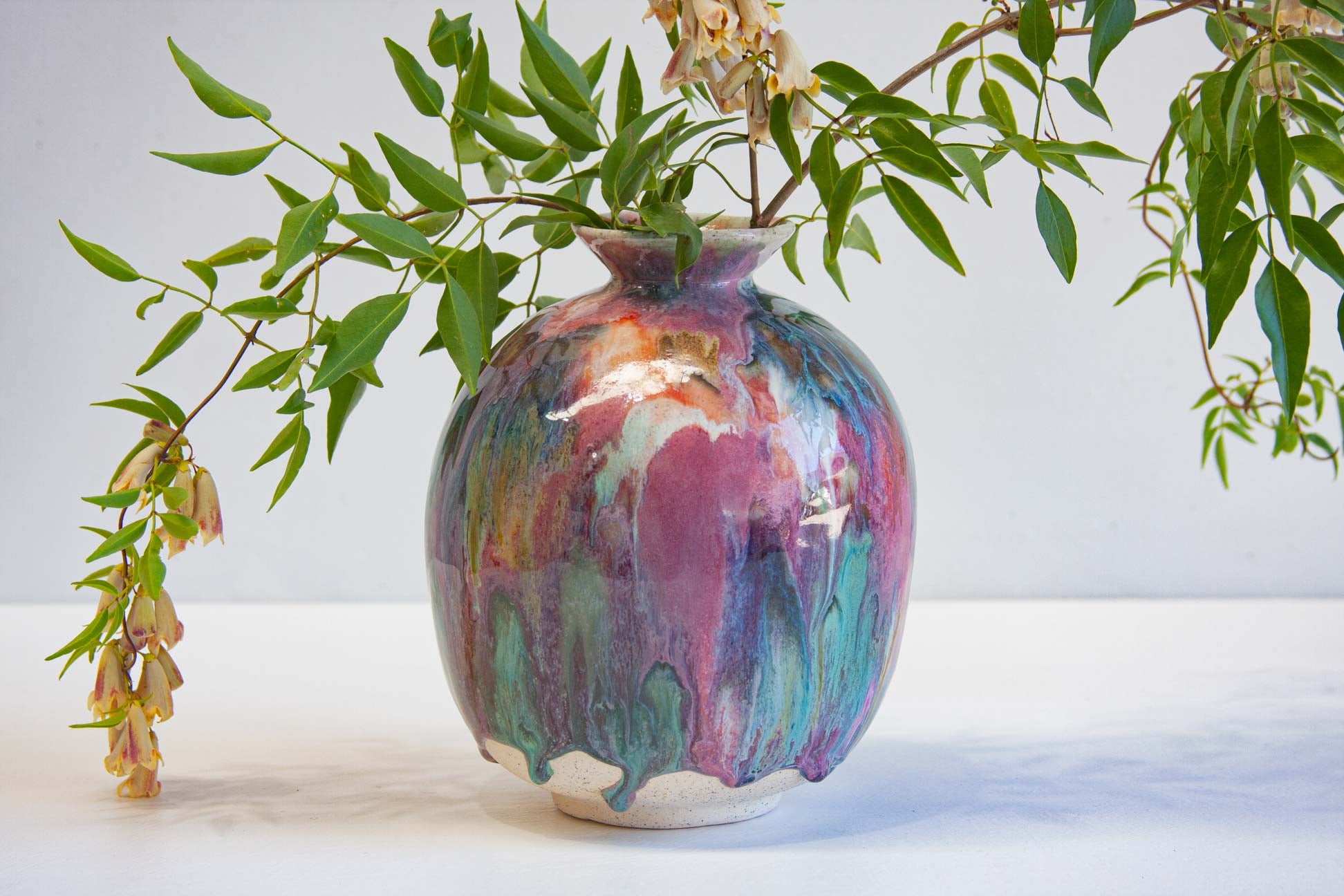 A ceramic vase with a glossy, dripping multicolor glaze in shades of purple, teal, orange, and cream, filled with green leafy branches against a plain white background.