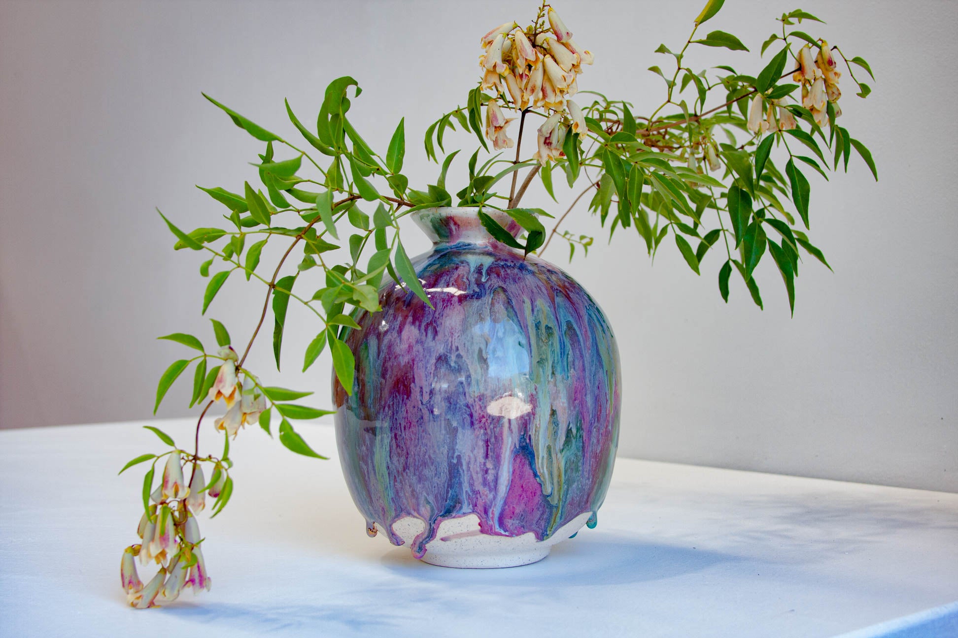 A ceramic vase with a marbled purple, blue, and white glaze, filled with green leafy branches and small cream flowers, set on a white surface against a light gray background.