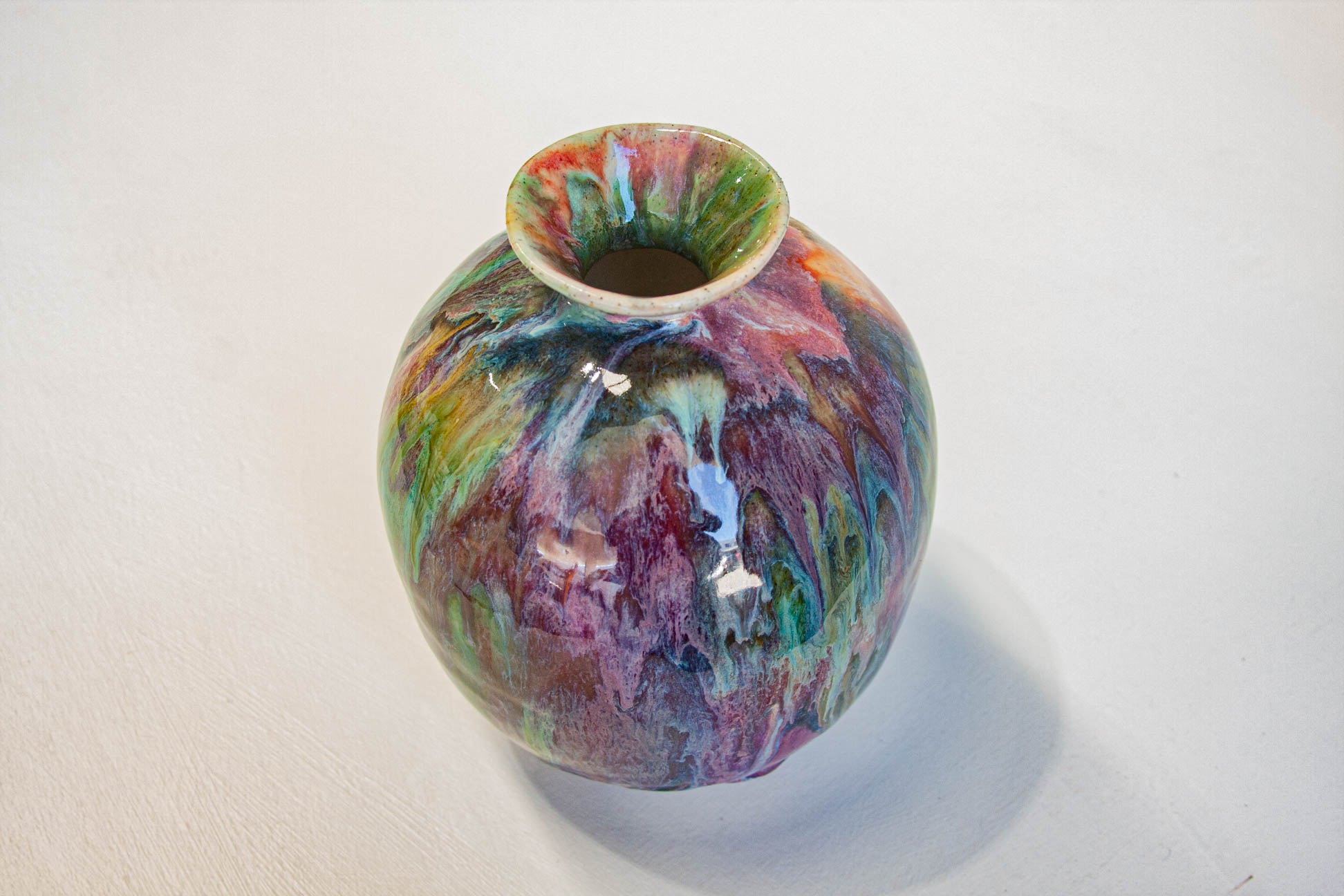 A small round ceramic vase with a flared rim, featuring a marbled, swirled glaze in multiple colors including purple, green, blue, orange, and pink, set against a plain off-white background.