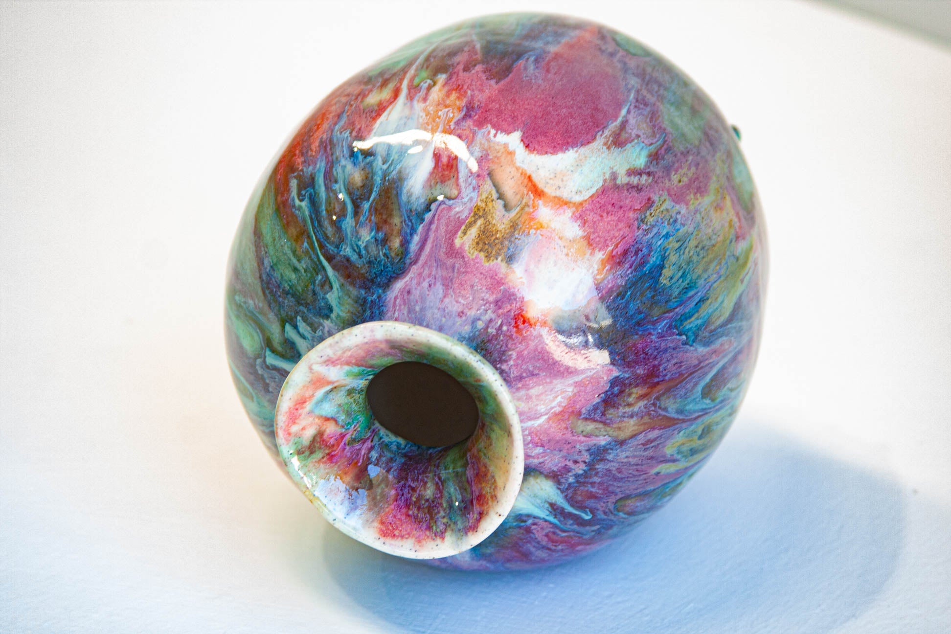 A small round ceramic vase with a narrow opening, decorated with a marbled swirl pattern of pink, purple, blue, green, and white glaze on a white background.