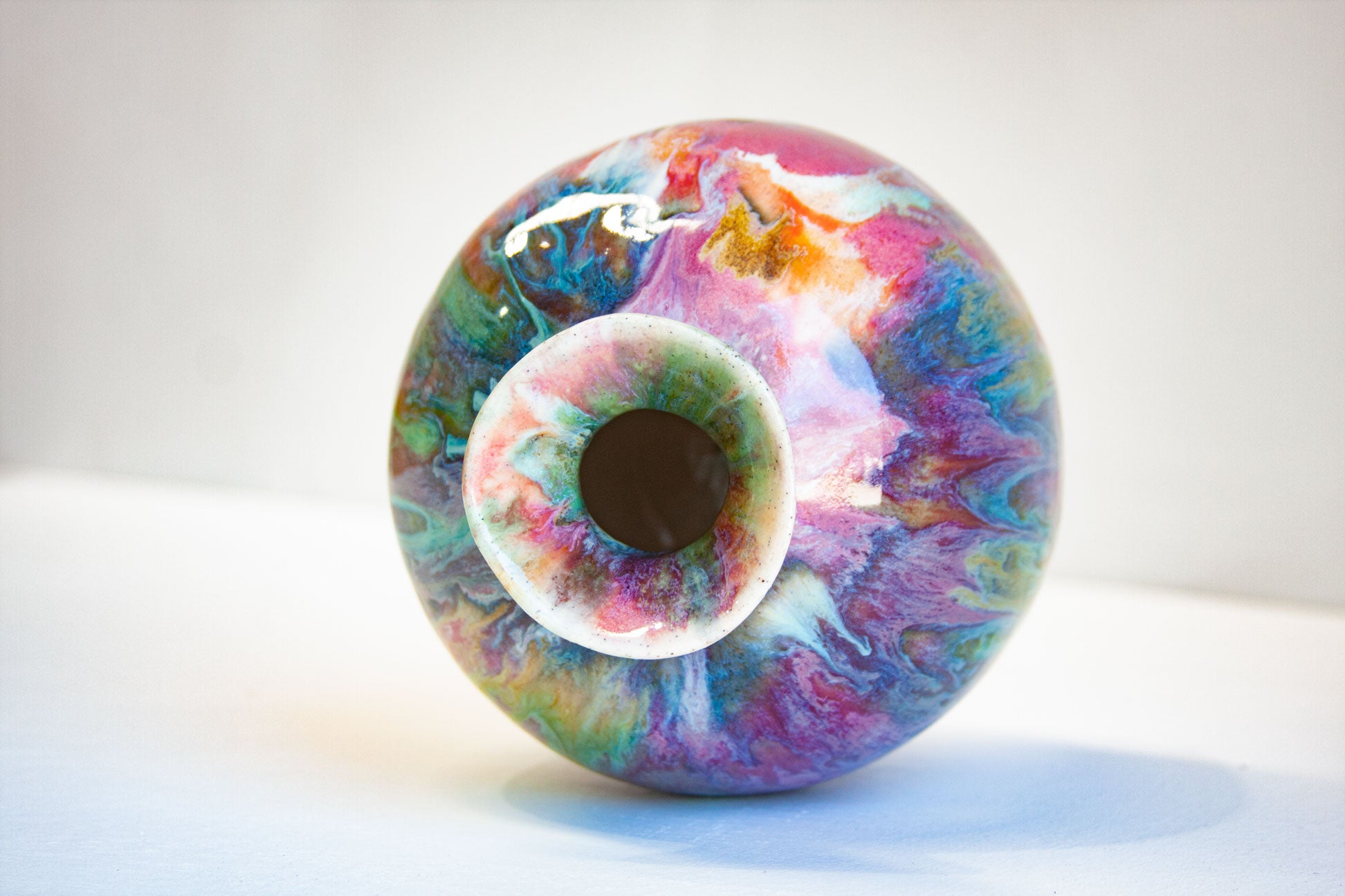 A round glass vase with a colorful marbled pattern of pink, purple, blue, green, and orange, featuring a wide flared rim and a dark interior opening, set against a plain light gray background.