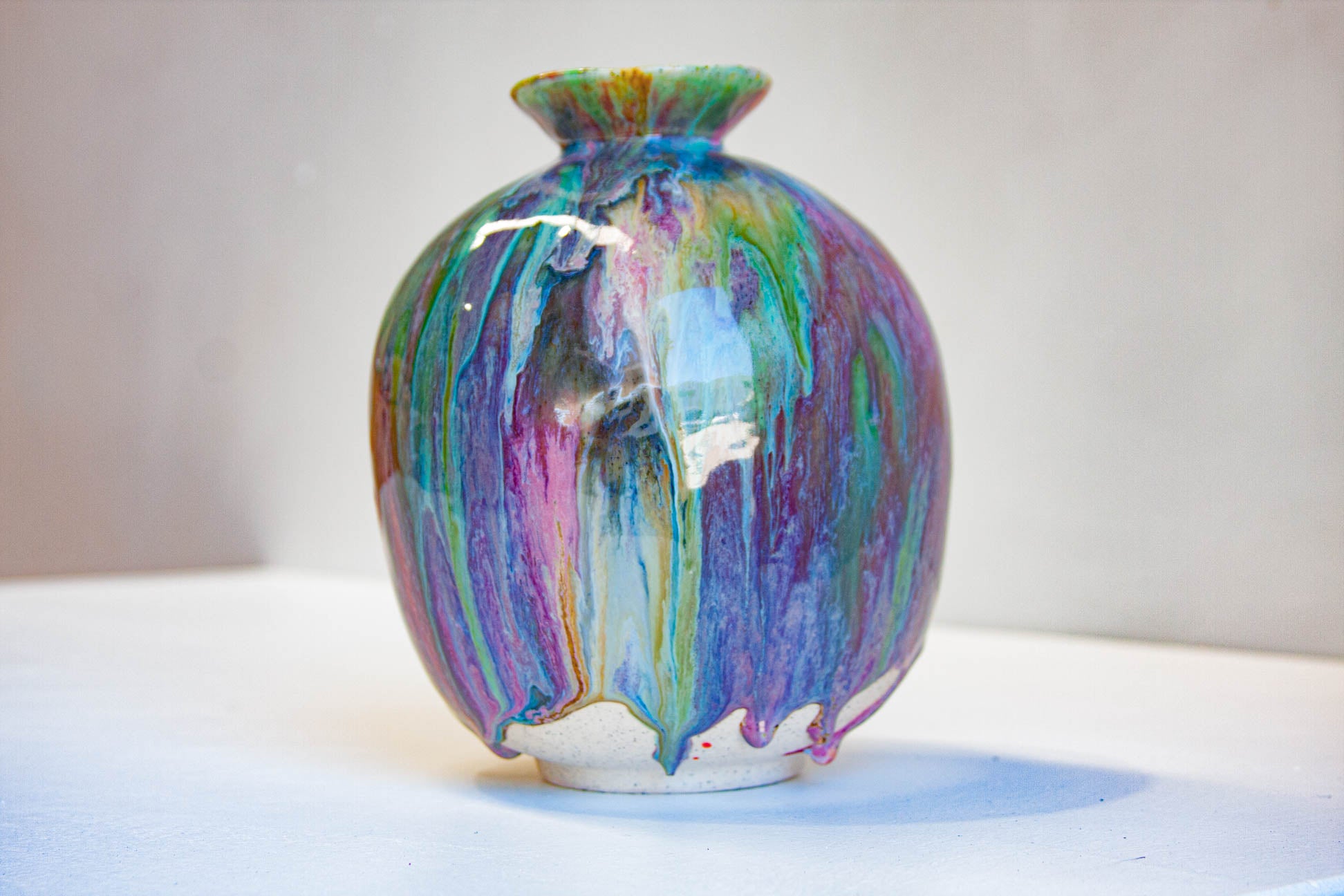 A small ceramic vase with a rounded body and short flared rim, covered in a glossy drip-glaze finish blending purple, blue, green, yellow, and orange tones, photographed against a plain light background.