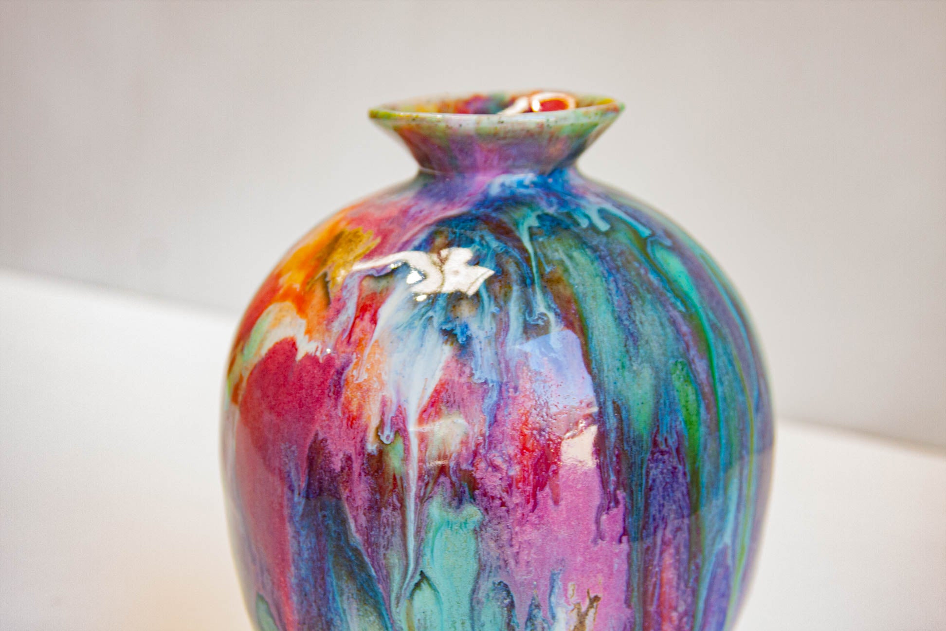 A small ceramic vase with a rounded body and narrow flared neck, decorated with a glossy multicolor drip glaze in pink, purple, blue, green, orange, and red tones, photographed against a plain light background.