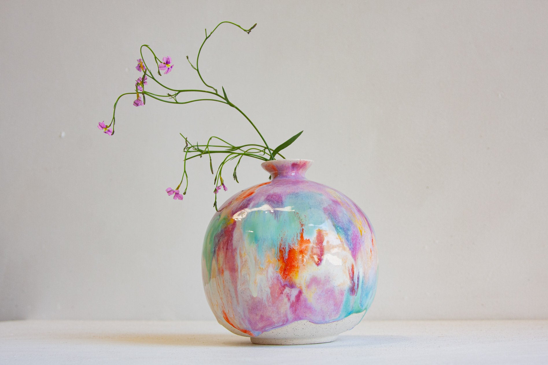 A round ceramic vase with a colorful marbled pattern in pink, purple, blue, and orange, holding delicate green stems with small pink flowers, set against a plain light gray background.